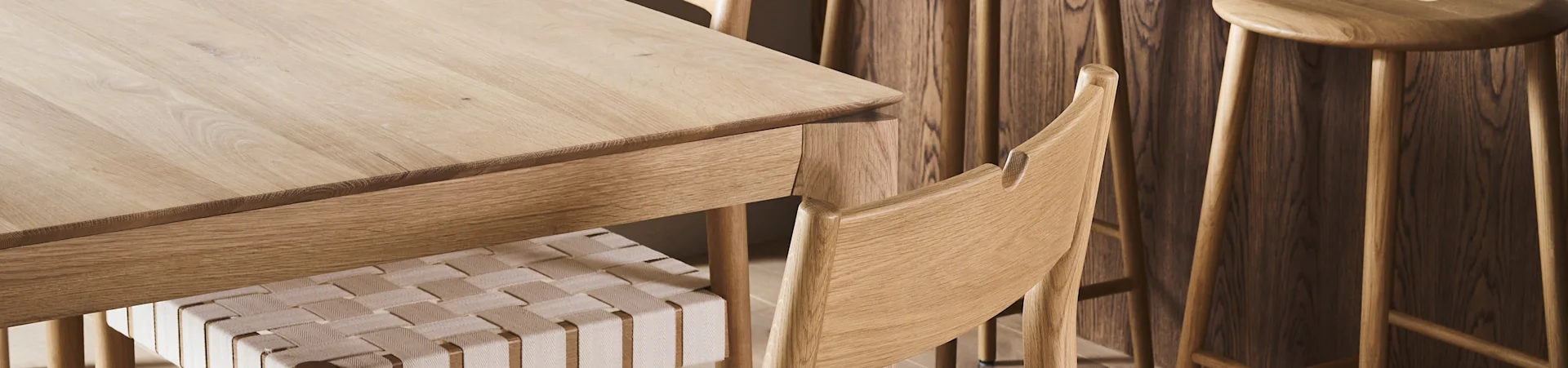 Nahaufnahme eines hellen Holzesstisches mit einem Stuhl mit geflochtener Sitzfläche und Holzschemeln im Hintergrund.
