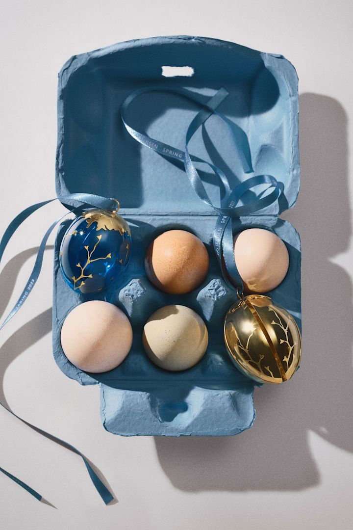 Das goldene und das blaue Dekorationsei aus der Spring 2025-Kollektion von Georg Jensen zusammen mit echten Eiern in einer Eierschachtel.