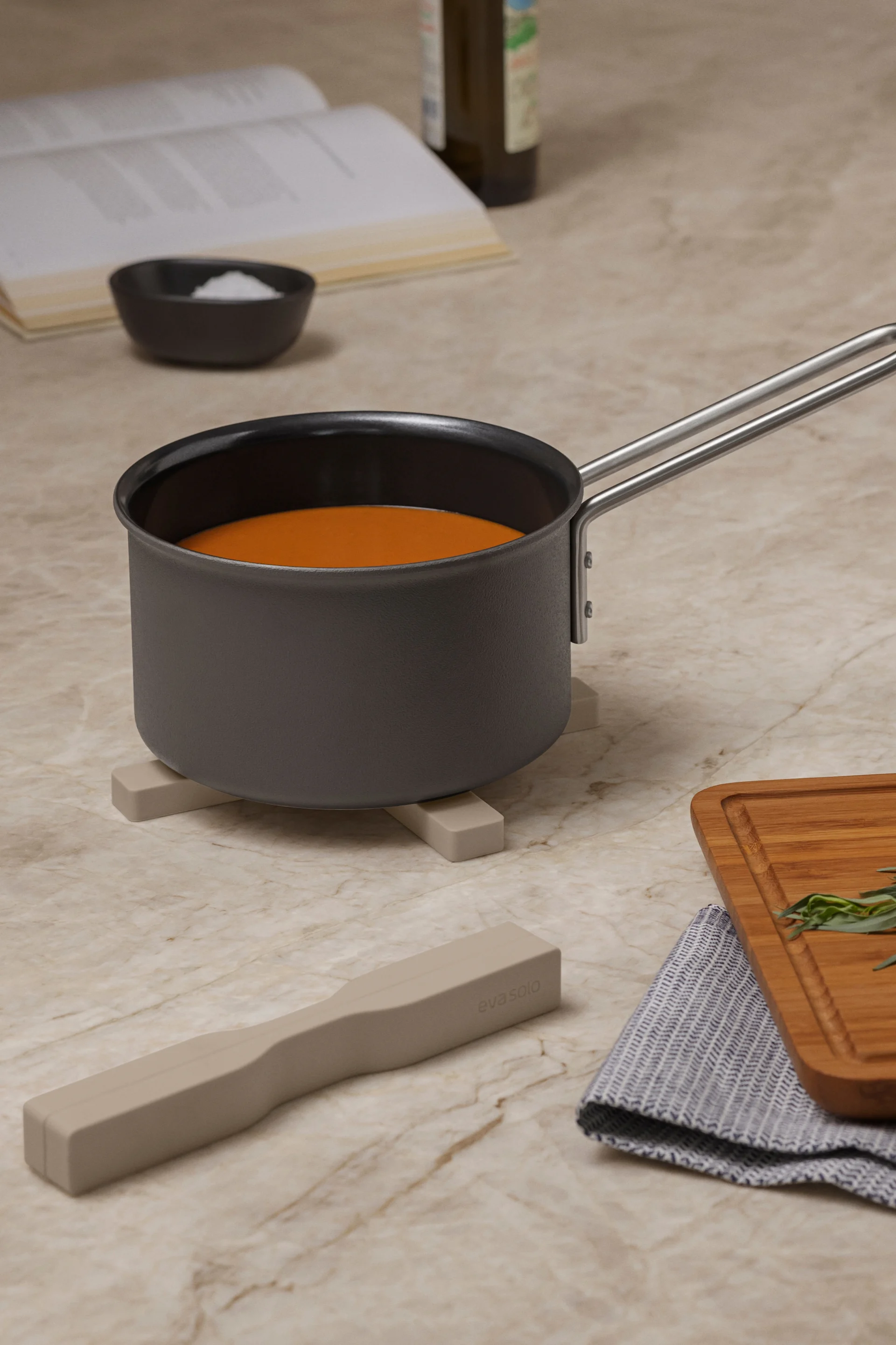 Topf mit oranger Flüssigkeit auf einem Untersetzer, dazu Kochbuch, Salz, Schneebesen, Kräuter auf Schneidebrett und Utensil auf Marmorplatte.