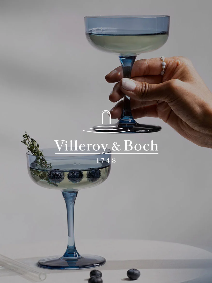 Hand hält blaues Coupe-Glas; ein weiteres Glas darunter mit Blaubeeren, Thymian und Villeroy & Boch Logo sowie '1748'.