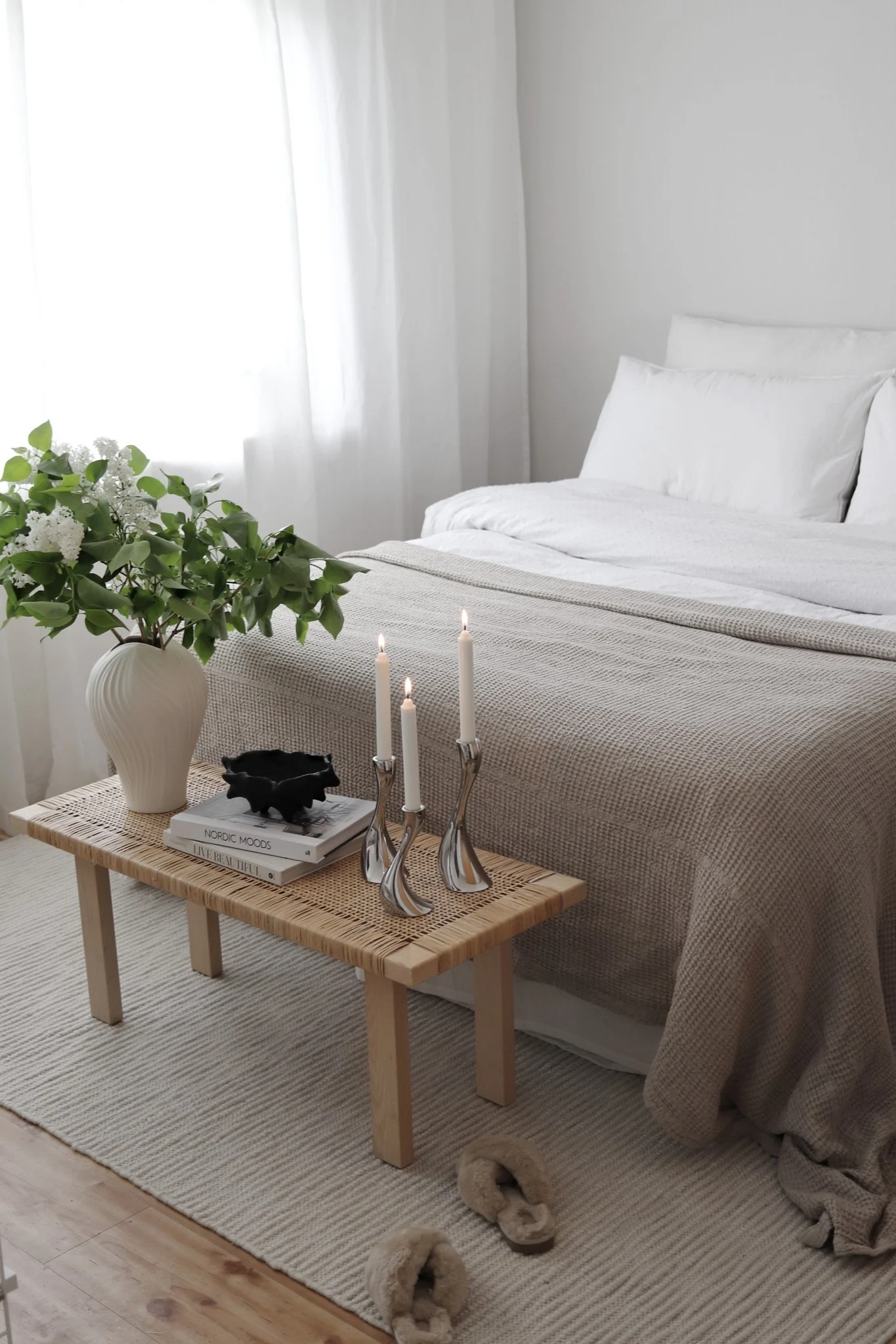 Teppiche fürs Schlafzimmer: Der weiße Pebble Wollteppich von Scandi Living liegt halb unter dem Bett, am Fußende steht eine kleine Bank, die mit einer Pflanze und Kerzen dekoriert ist. @hemmahosfalk