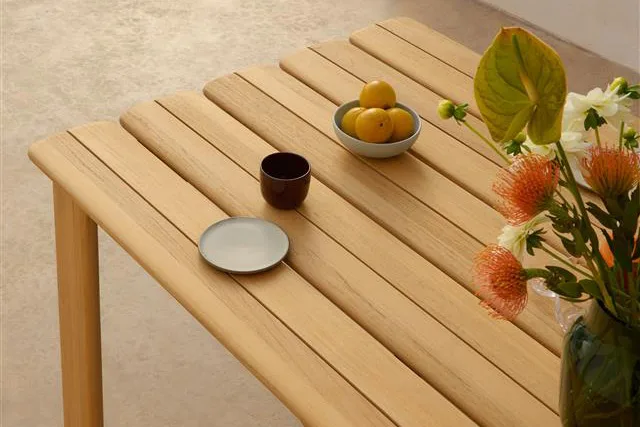 Heller Holztisch mit Lamellen, einer Schale Zitronen, dunkler Tasse, kleinem Teller und einer Vase mit orange-weißen Blumen.