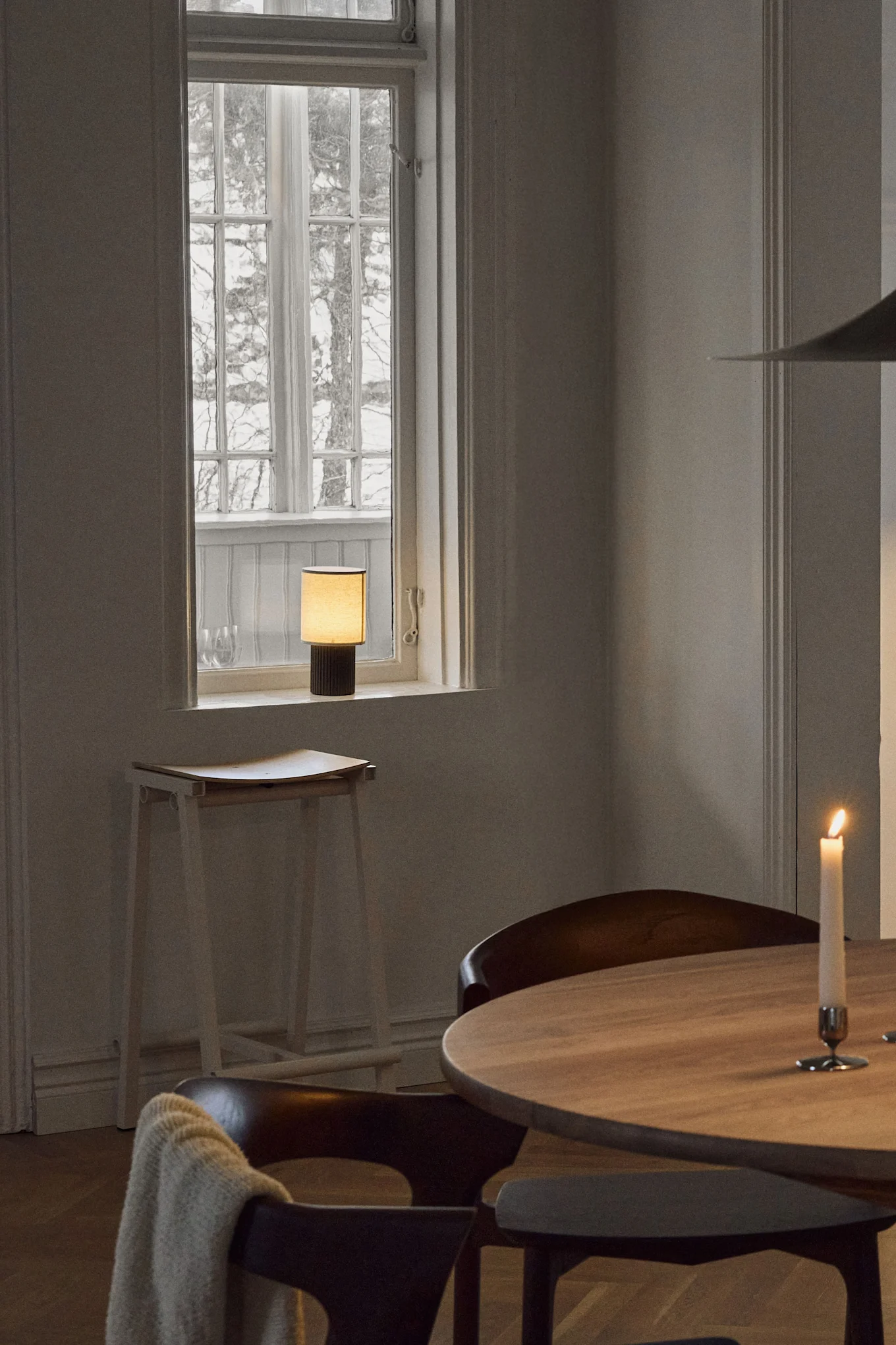 Ein gemütlicher Raum mit einer Akku-Tischleuchte auf der Fensterbank, einem runden Holztisch und einer brennenden Kerze.