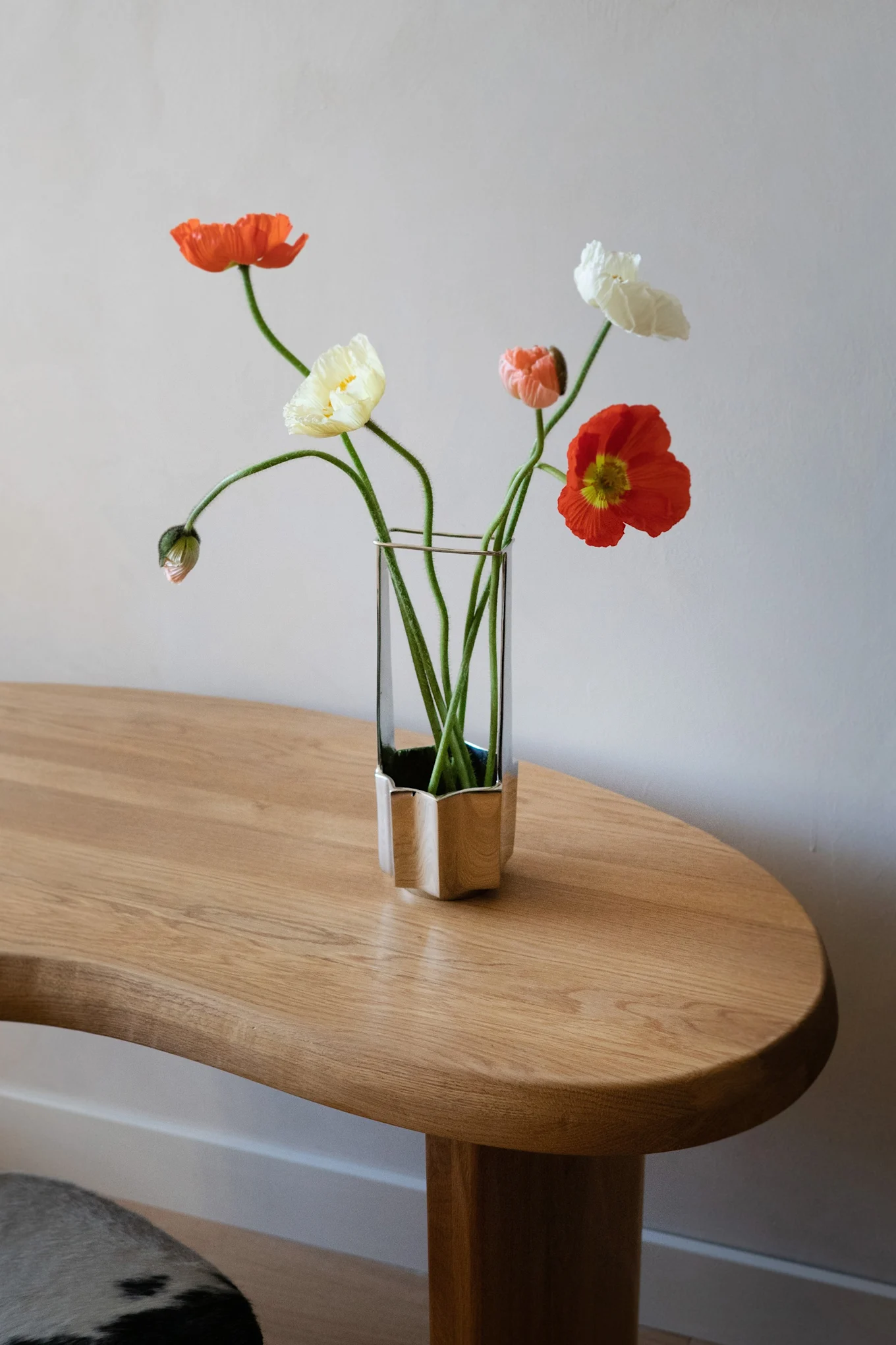 Ein Strauß orangefarbener, gelber und weißer Mohnblumen mit langen grünen Stielen in einer einzigartigen Glas- und Silbervase auf einem hellen Holztisch.
