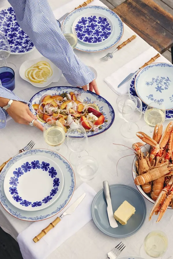 Person platziert eine Platte mit Pfirsich-Feta-Salat auf einem Tisch mit blau-weißem Geschirr, Wein und Langusten.