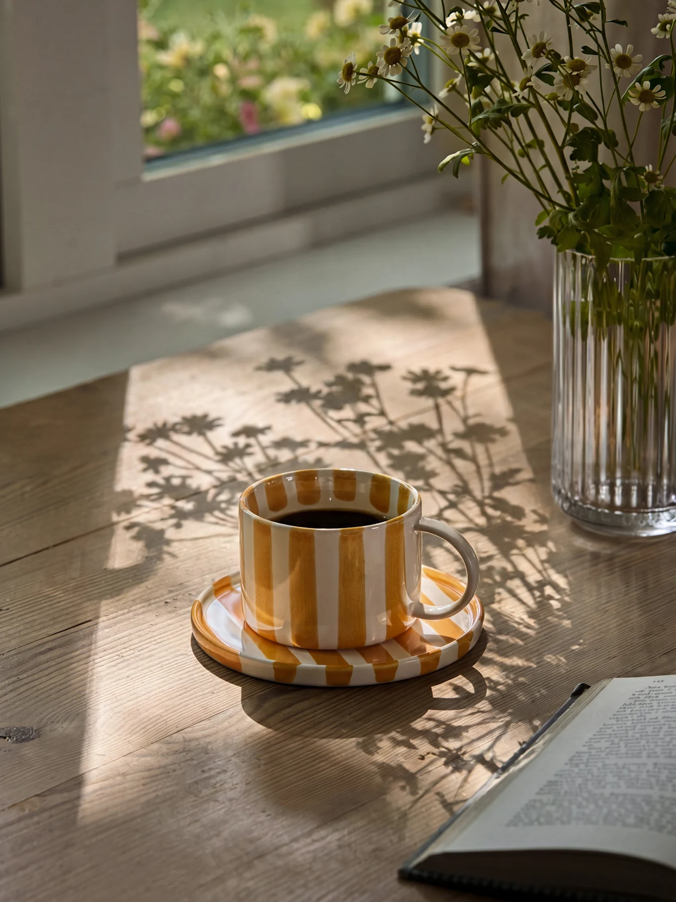 Gelb-weiß gestreifte Kaffeetasse mit schwarzem Kaffee auf einem Holztisch mit Sonnenschatten und einer Vase mit Gänseblümchen.