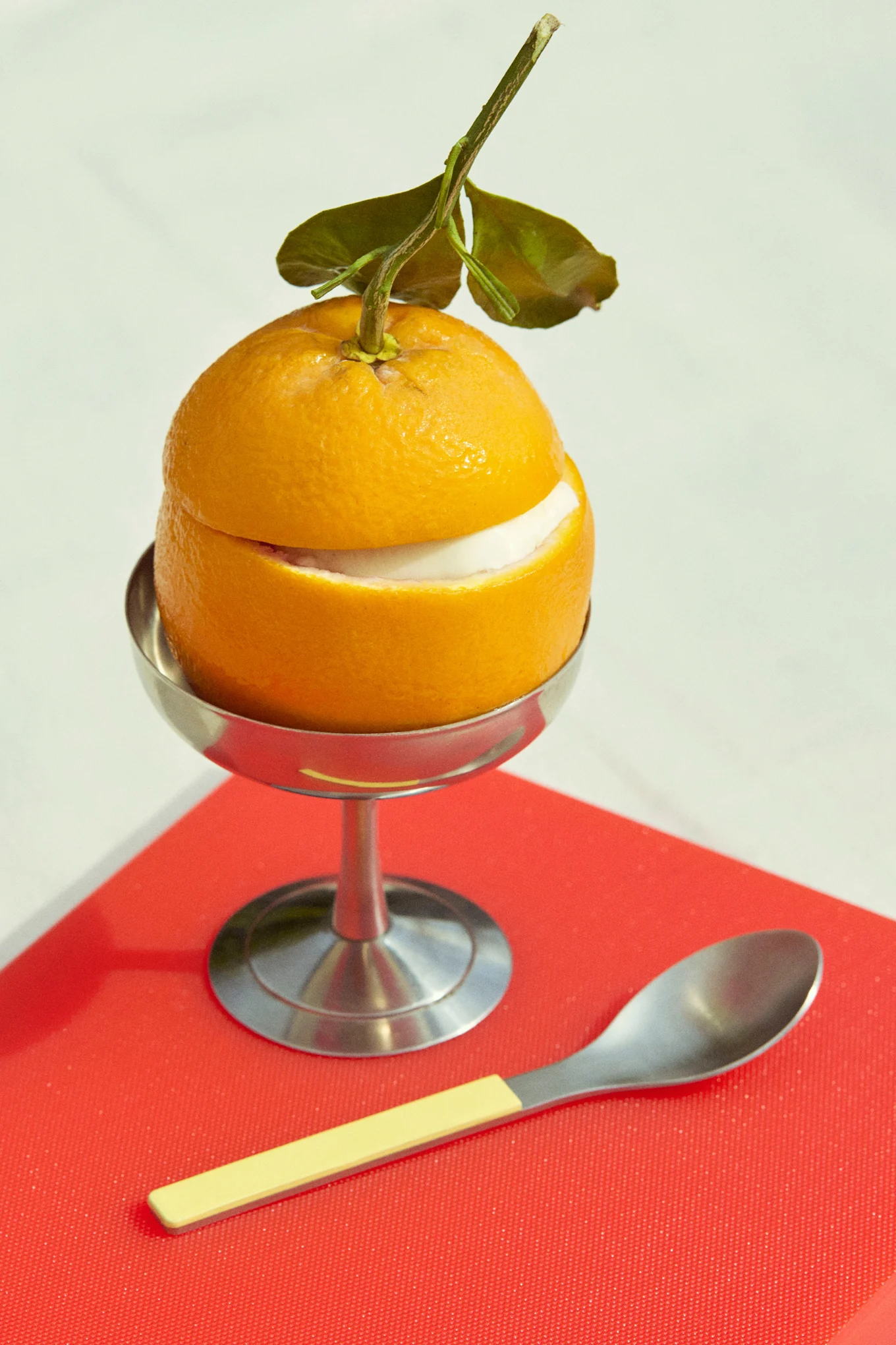 Orangendessert mit weißer Füllung in einem Silberbecher, mit Löffel, auf roter Oberfläche.