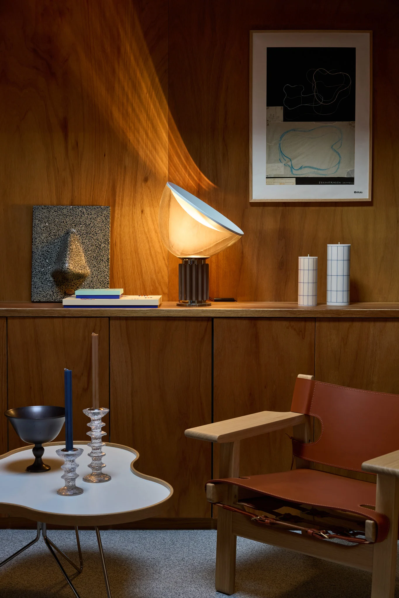 Eine Lampe beleuchtet ein holzvertäfeltes Zimmer mit Regalen, Kunst, Kerzen, einem Tisch und Ledersessel.