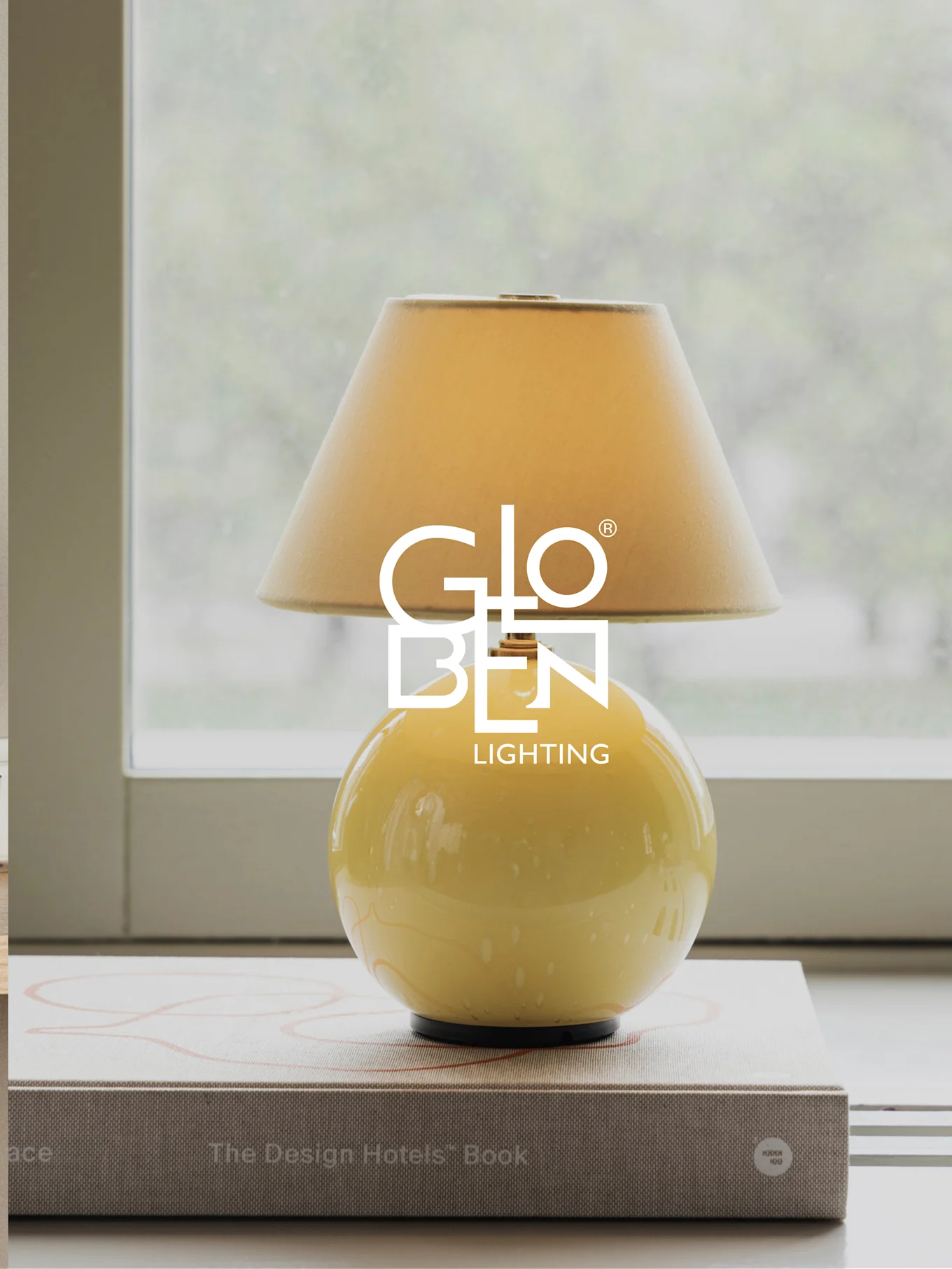 Gelbe Tischlampe mit beigem Schirm, steht auf Büchern an einem Fenster.