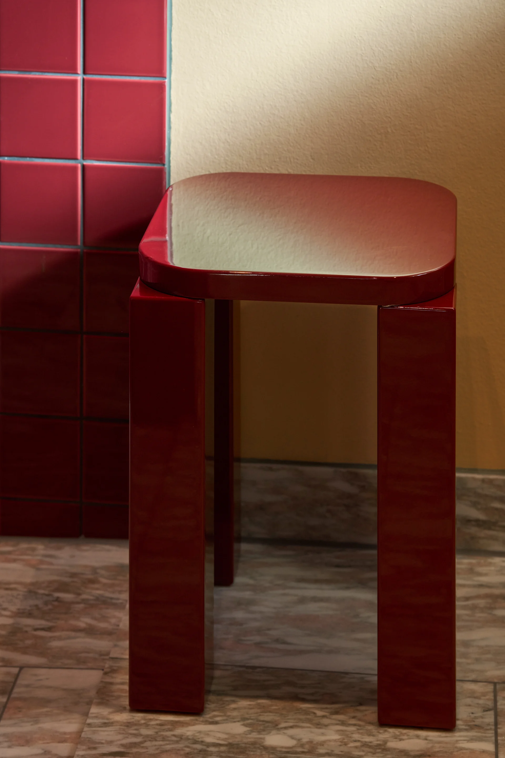 Ein glänzender, dunkelroter Hocker mit rechteckiger Platte und vier Beinen steht auf einem Marmorboden neben einer gefliesten Wand.