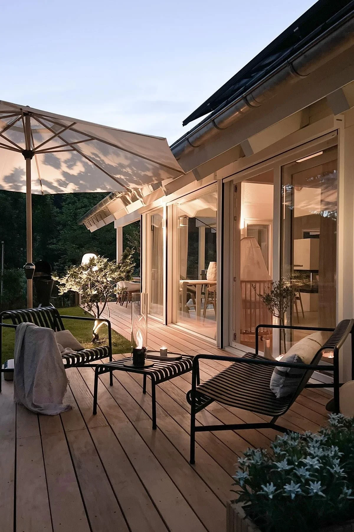 Gemütliche Holzterrasse in der Abenddämmerung mit zwei schwarzen Latten-Loungesesseln, einem Couchtisch mit Kerze und einem modernen Haus mit beleuchteten Innenräumen.