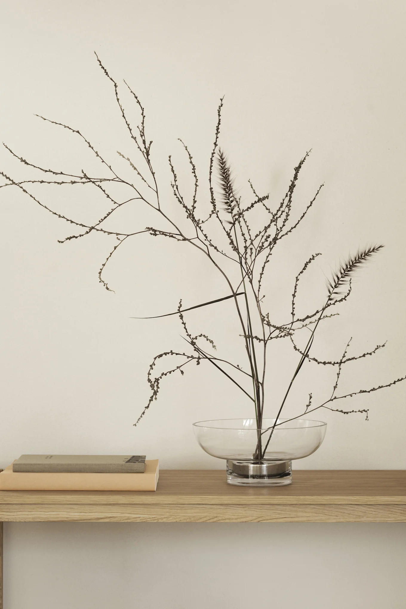 Ein minimalistisches Arrangement auf einem Holzregal mit zwei gestapelten Büchern und einer Glasschale mit getrockneten Zweigen und Federgras vor einer hellen Wand.