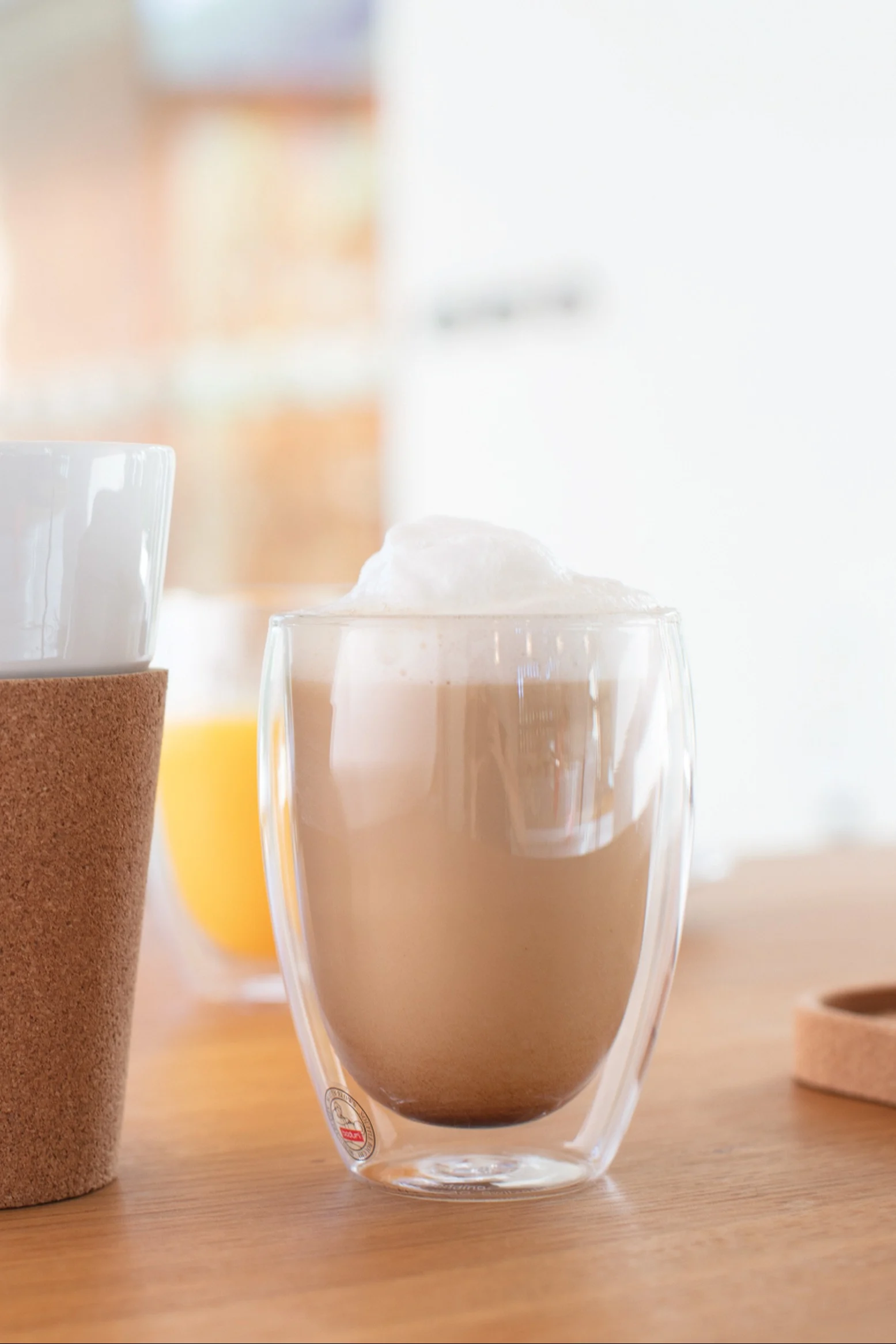 Schaumiger Latte in einer doppelwandigen Klarglastasse auf einem Holztisch, links daneben eine weitere Tasse mit Korkmanschette und ein oranges Getränk im Hintergrund.
