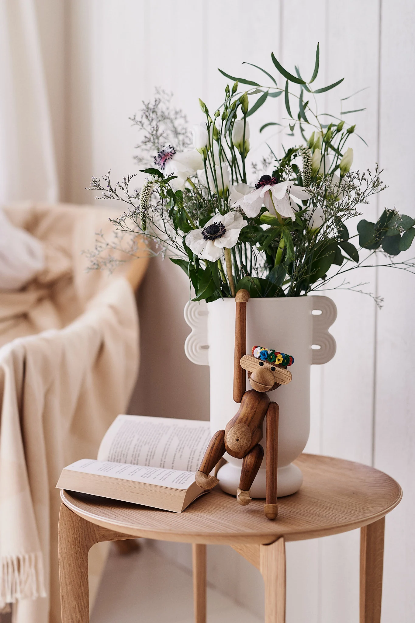 Ein Holzaffen-Spielzeug hängt an einer weißen Vase mit weißen Blumen neben einem offenen Buch auf einem Holztisch.