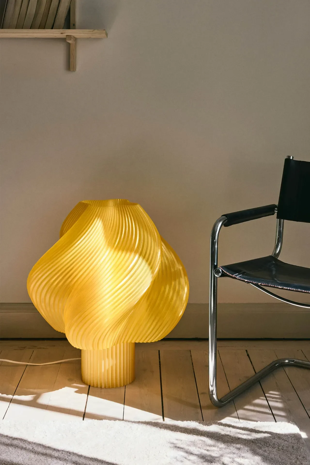 Eine hellgelbe, skulpturale Tischlampe mit geripptem, welligem Design leuchtet auf einem Holzfußboden neben einem schwarz-ledernen Chromstuhl.