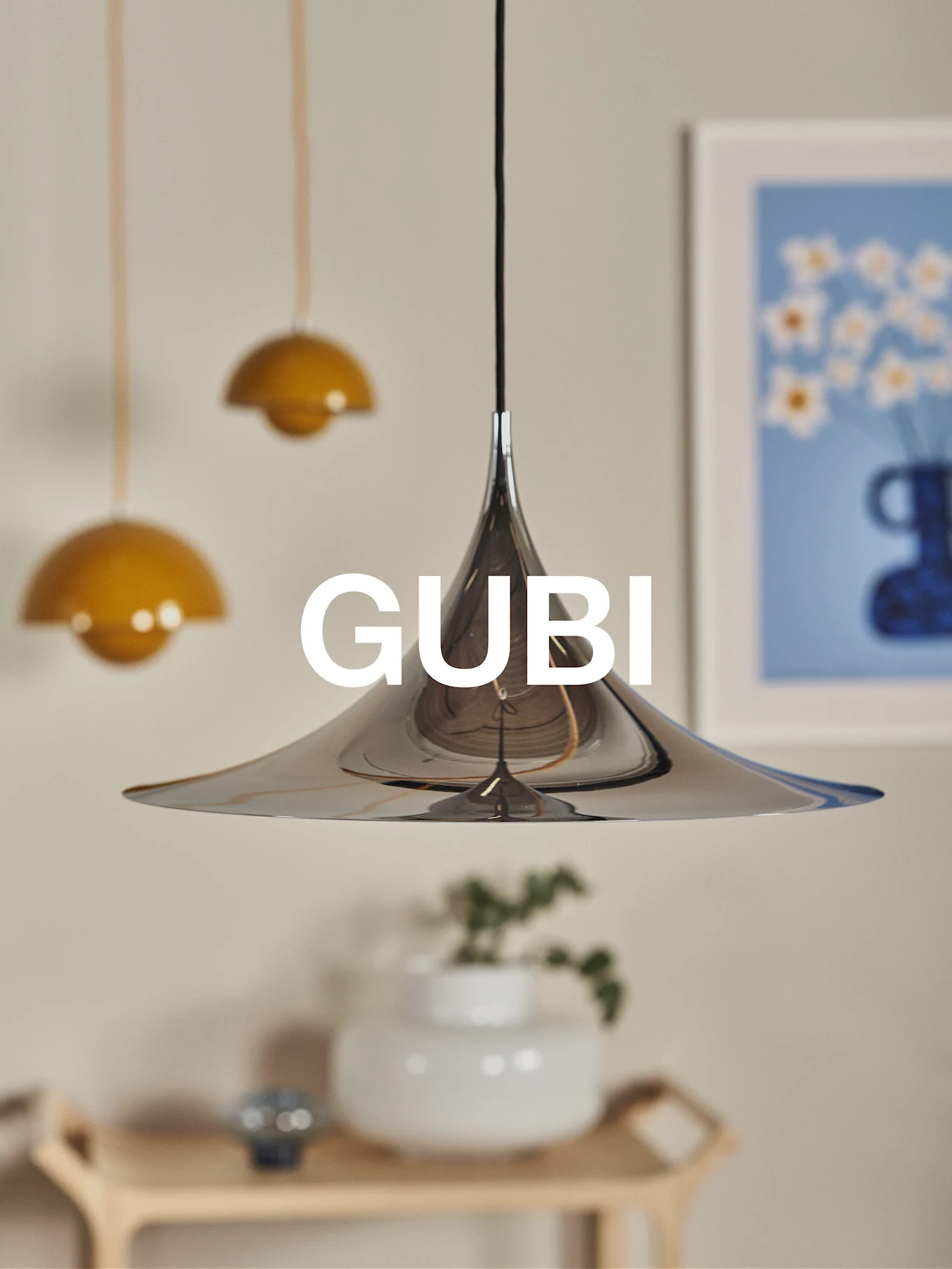 Eine polierte konische Chrom-Pendelleuchte mit GUBI-Logo vor verschwommenem Hintergrund.