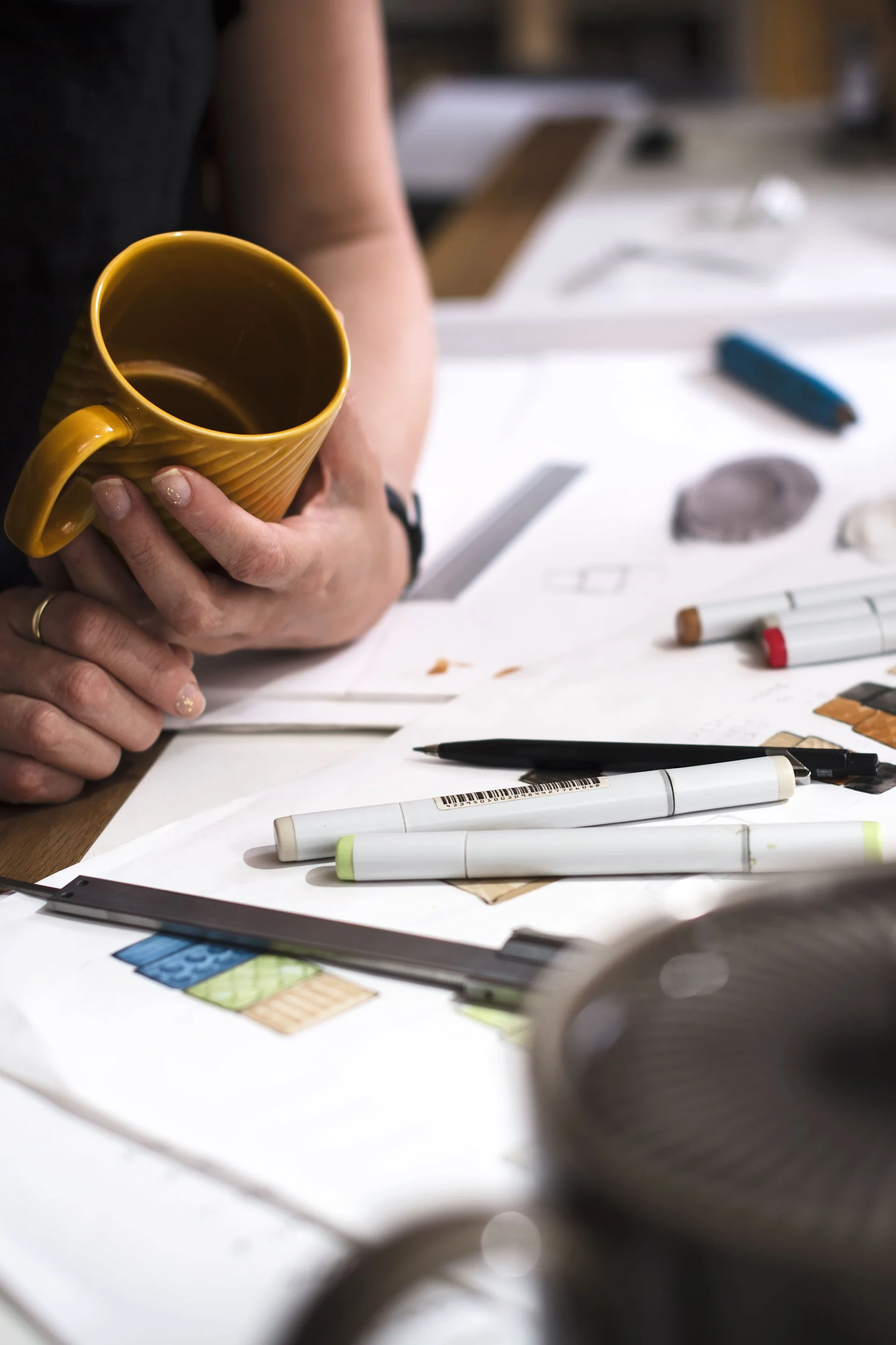 Ein Tisch mit Papier und Stiften, daneben steht eine Person mit dem gelben Kaffeebecher aus der Coffee & more-Kollektion von Sagaform in der Hand.