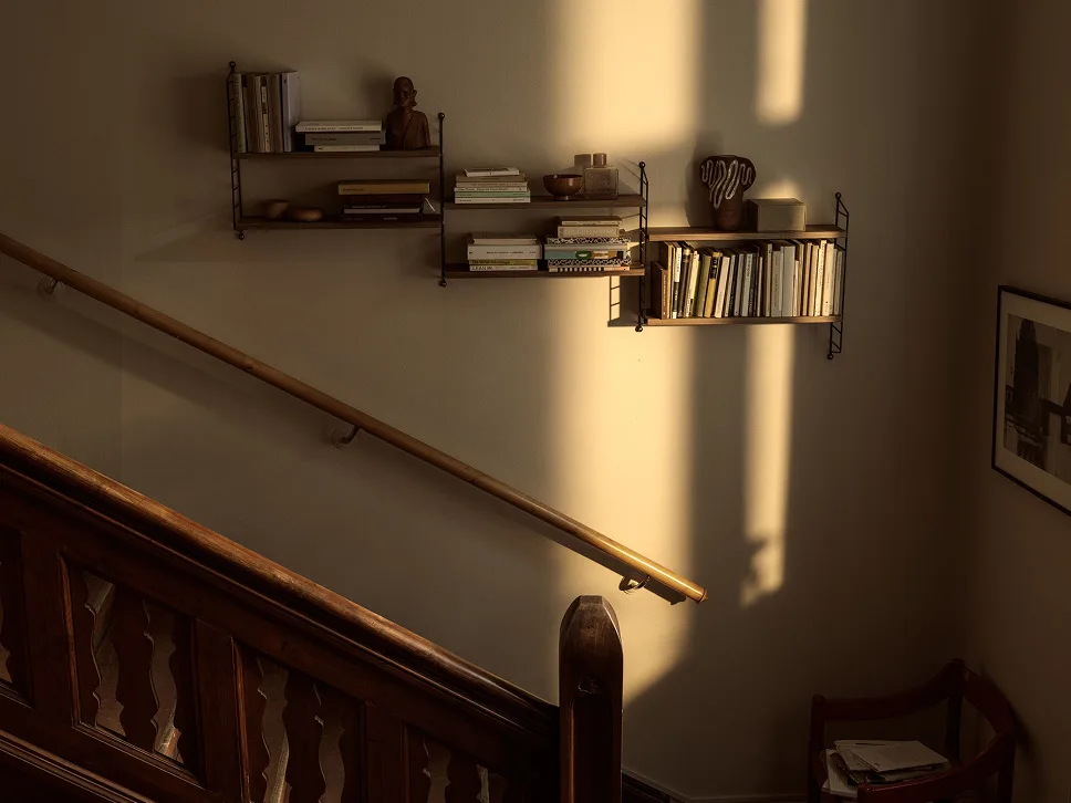 Sonnenbeschienene Wand mit Holzregalen voller Bücher und Dekoration, daneben eine Holztreppe mit Geländer.