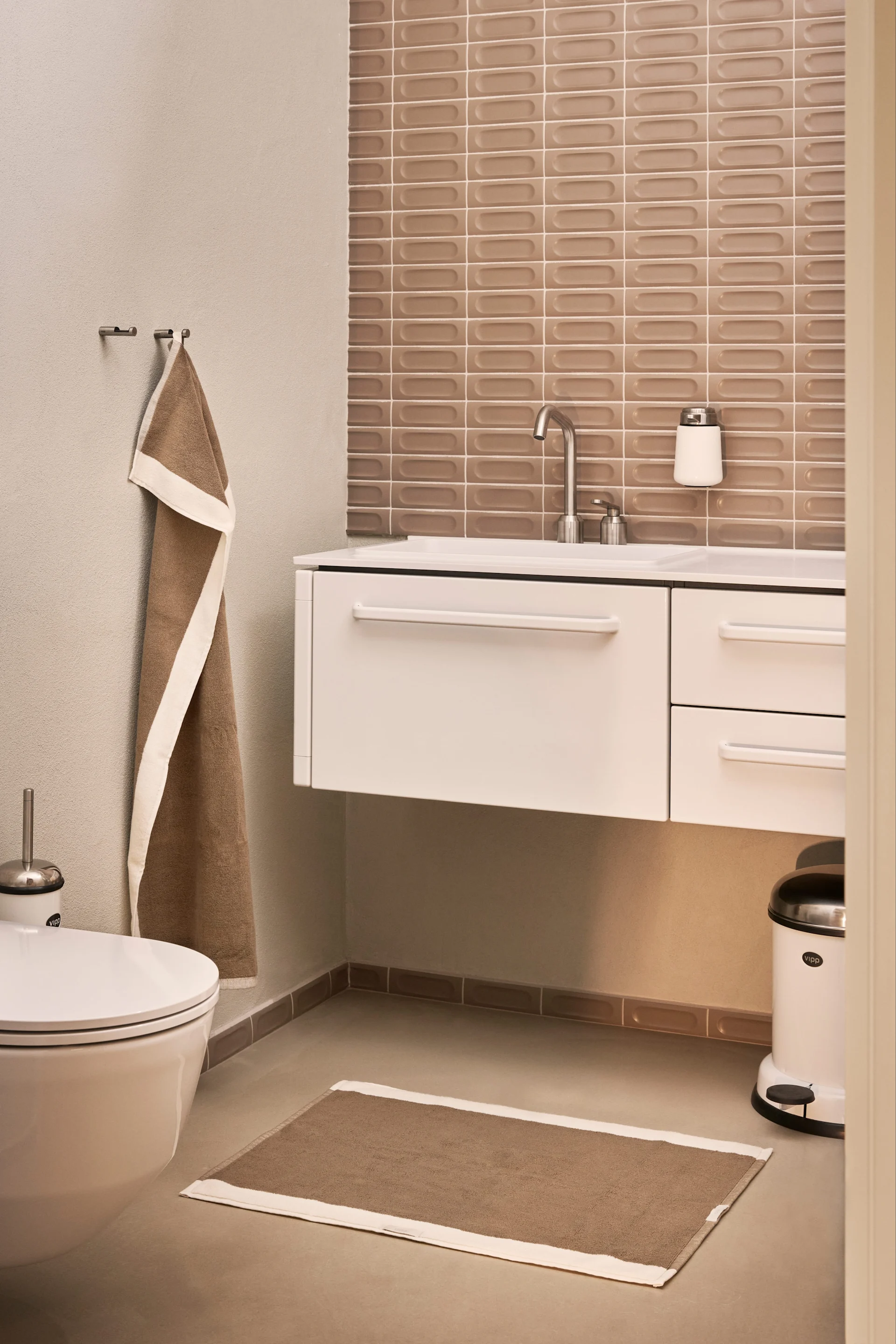 Ein modernes Badezimmer mit einem schwebenden weißen Waschtisch und Waschbecken, einer Toilette, einem braunen Handtuch und einer passenden Badematte.