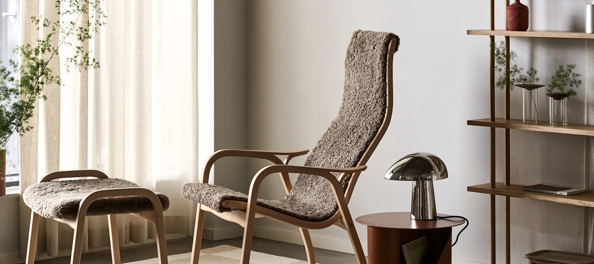 Heller Holz-Sessel und passender Hocker mit texturiertem braunem Bezug, eine metallische Tischlampe und ein Holzregal mit Pflanzen.