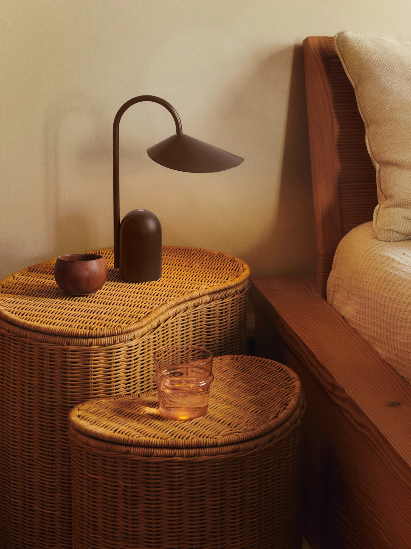 Zwei geflochtene Rattan-Nachttische mit einer dunkelbraunen Lampe, einer Holzschale und einem Glas Wasser.