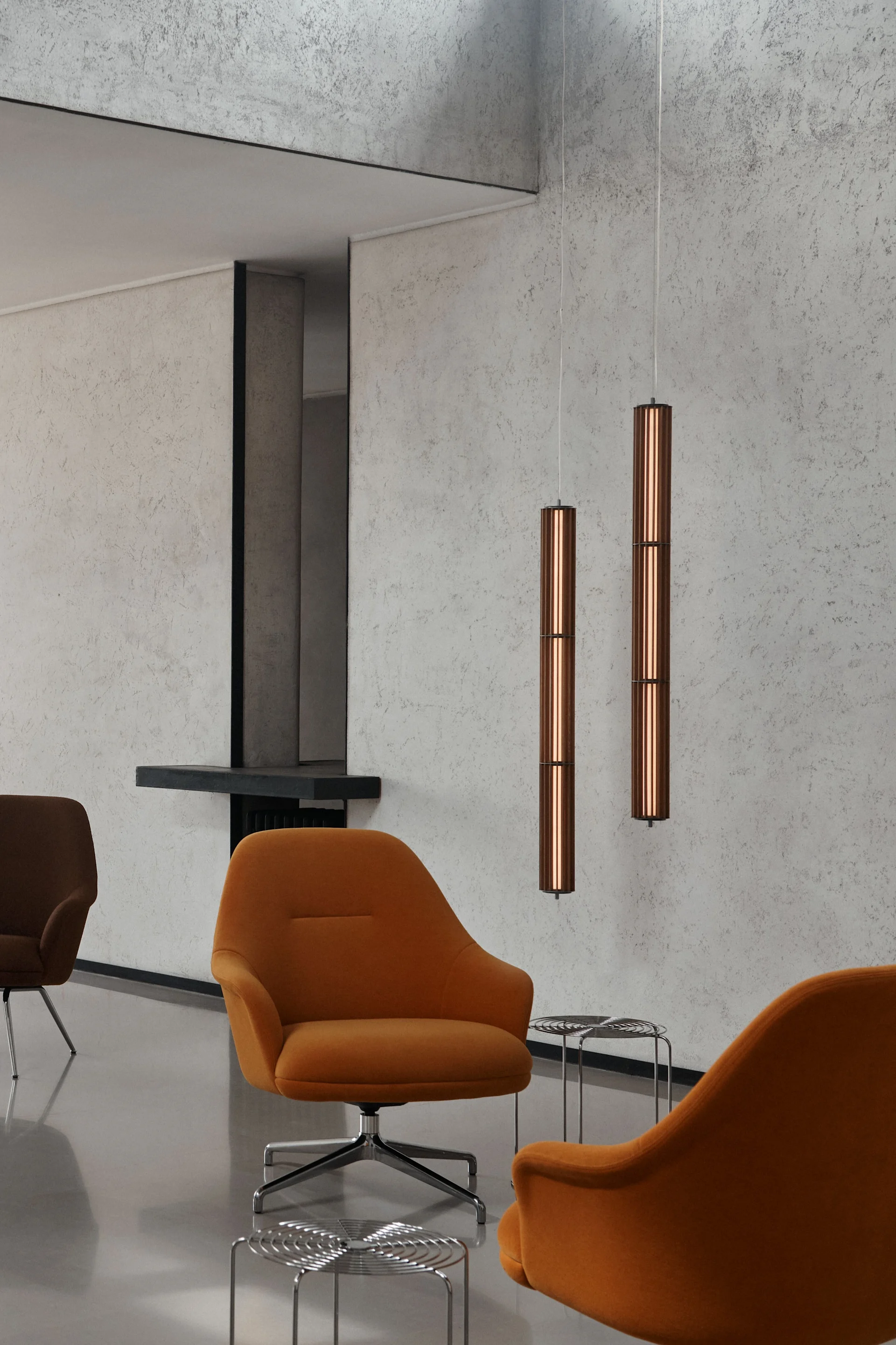 Zwei zylindrische Kupfer-Pendelleuchten hängen in einem modernen Raum mit orangefarbenen Sesseln und Wänden mit Betontextur.