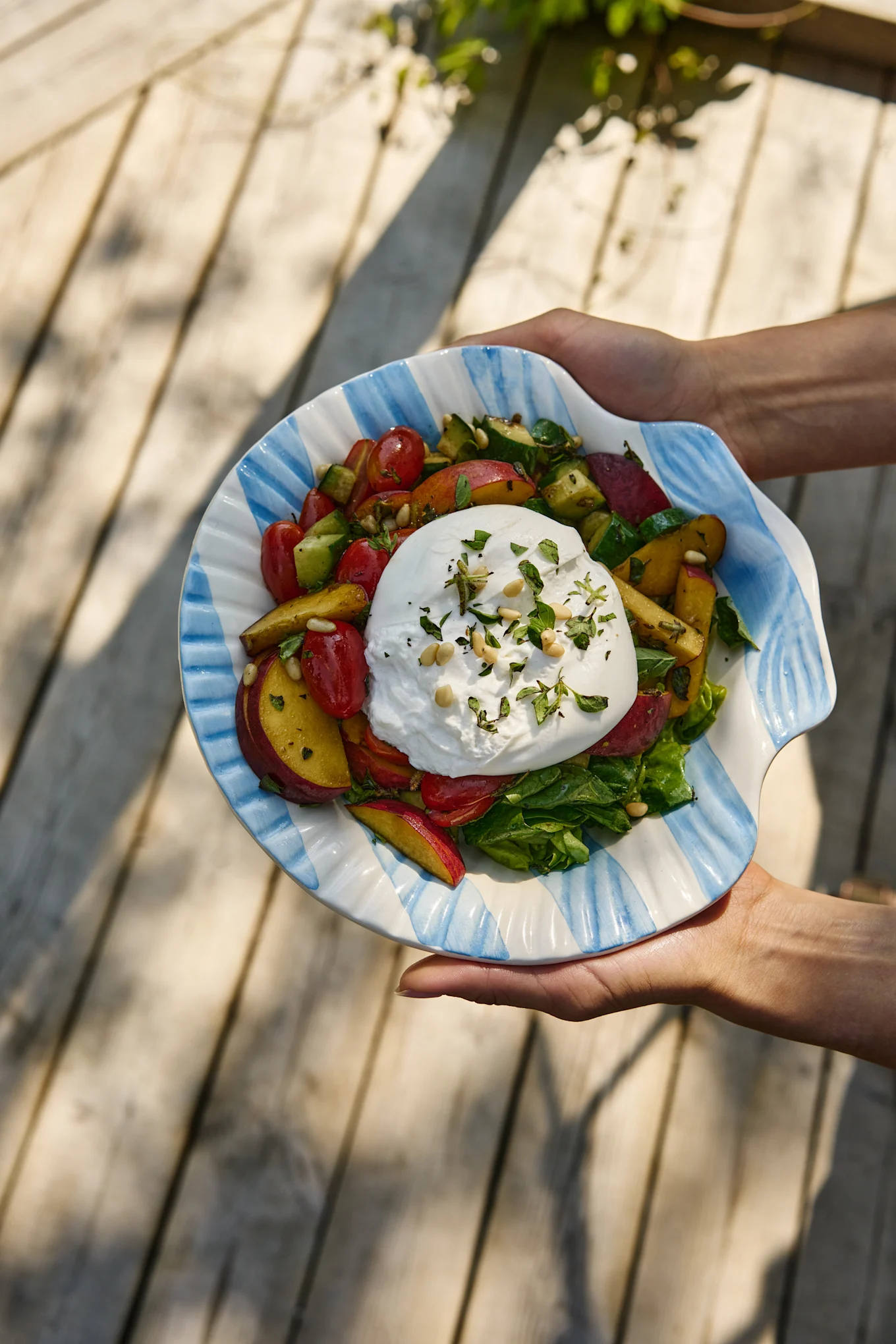 Hände halten einen Burrata-Salat mit Tomaten, Pfirsichen, Gurken und Grünzeug in einer gestreiften Muschelschale.