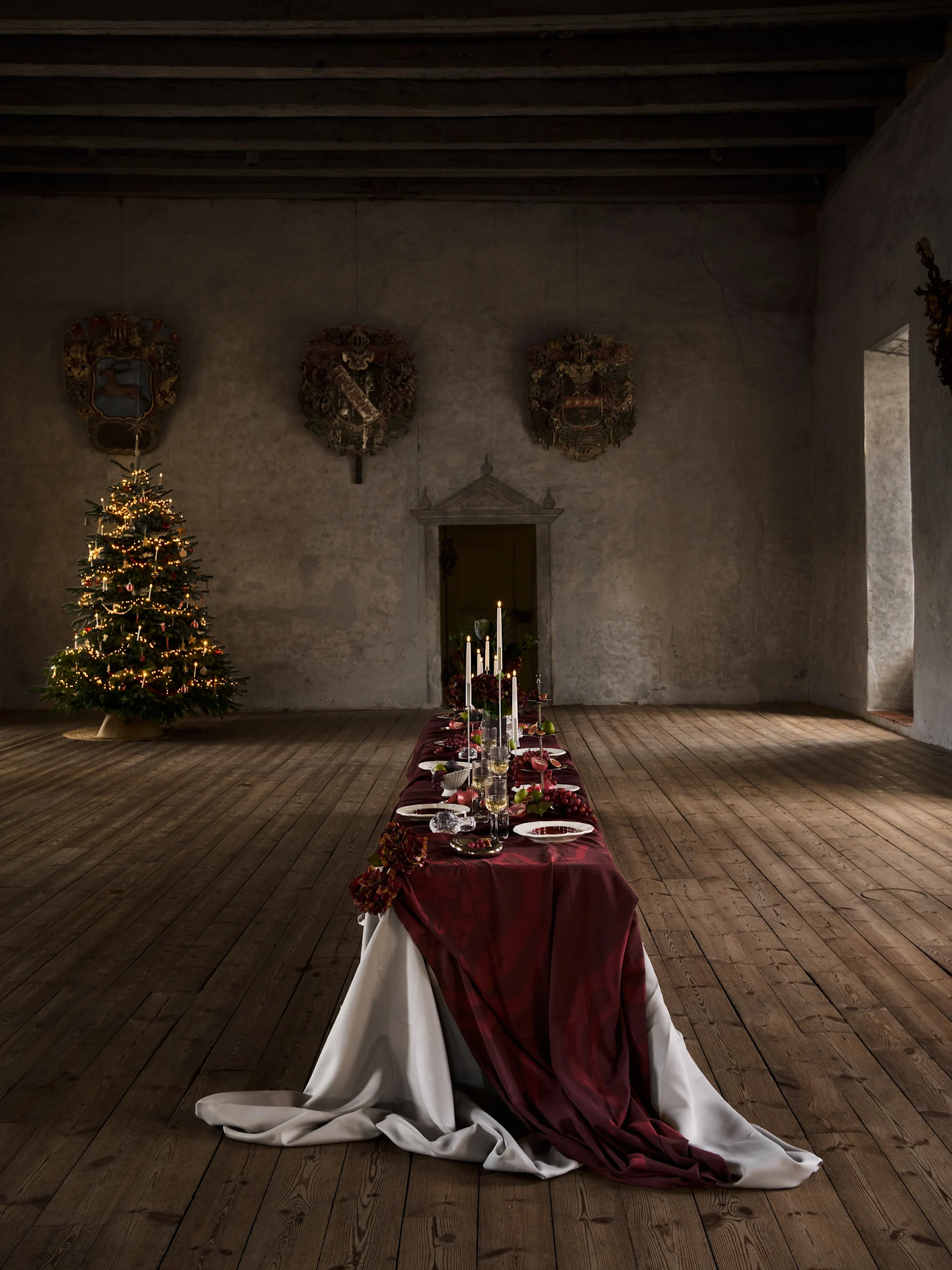 Langer Weihnachtstisch mit roter Decke, Kerzen, Obst und Gedeck, neben einem beleuchteten Weihnachtsbaum in einer alten Halle.