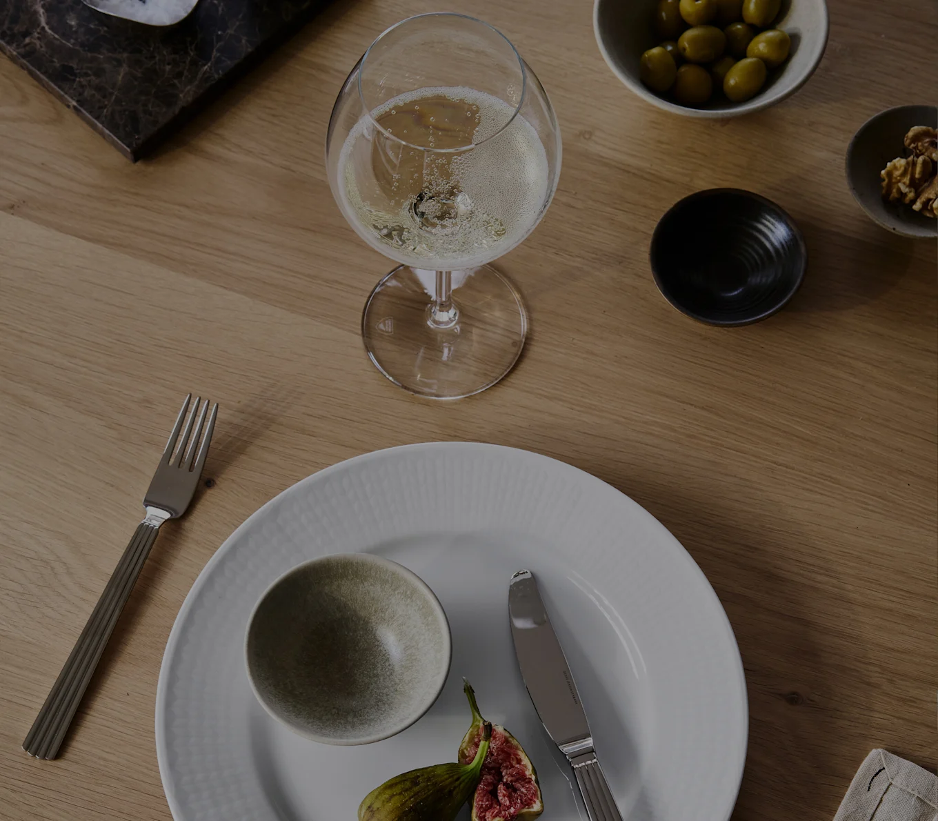 Ein Glas Champagner, weißer Teller mit Schale und Feigen, Gabel, Messer, Oliven und Walnüsse auf einem Holztisch.