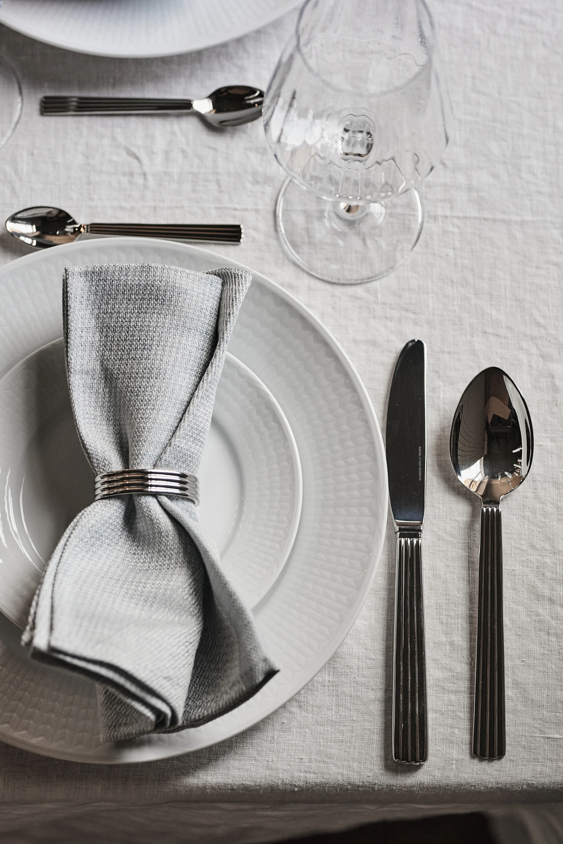 Elegante Tischdekoration mit weißen Tellern, einer grauen Serviette in einem Silberring und geripptem Besteck auf einem hellen Leinentischtuch.