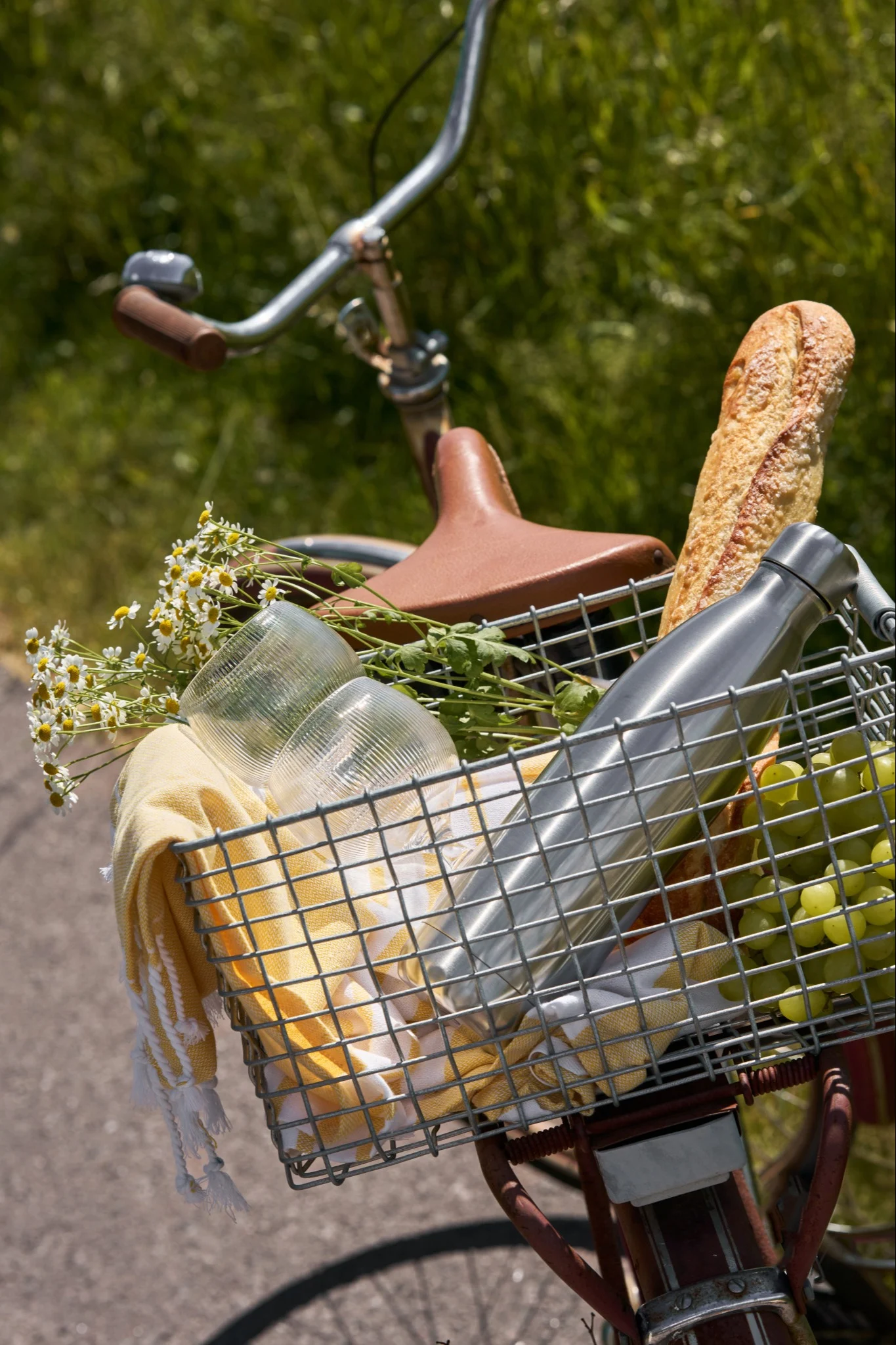 Fahrrad mit einem Vorderkorb, gefüllt mit Picknickartikeln: Brot, grüne Trauben, eine silberne Wasserflasche, Becher, ein gelbes Handtuch und Blumen.