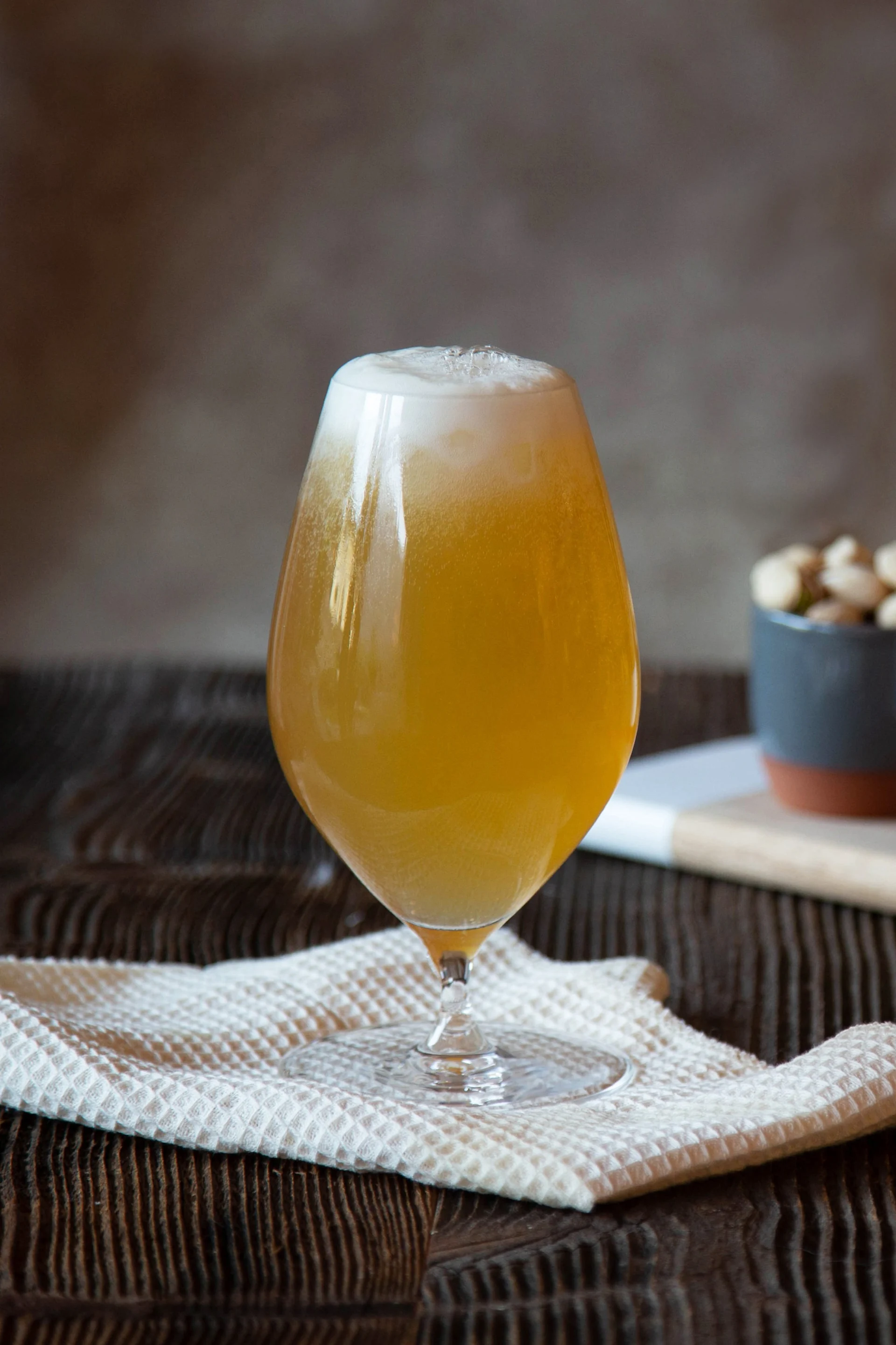 Ein Glas schaumiges bernsteinfarbenes Bier auf einem weißen Tuch, mit Schalen voller Nüsse und Grissini im Hintergrund auf einem Holzbrett.
