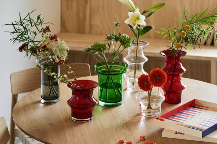 Eine Kollektion von roten, grünen, grauen und durchsichtigen Vasen stehen auf einem Tisch neben einem rot-blauen Backgammon-Spiel.