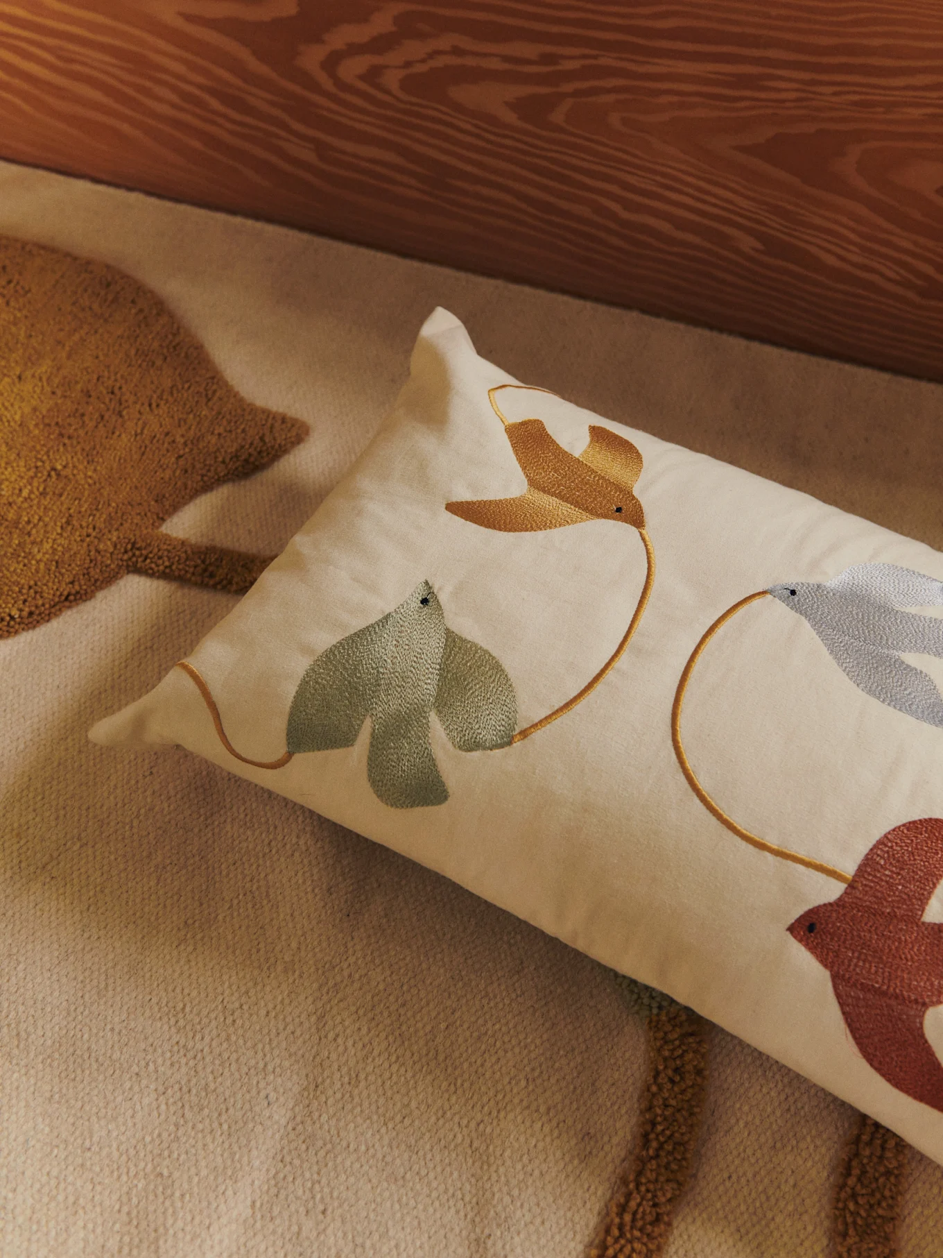 Lendenkissen mit gestickten Vögeln und Blättern in Gold, Grün, Blau und Rotbraun auf hellem Teppich.