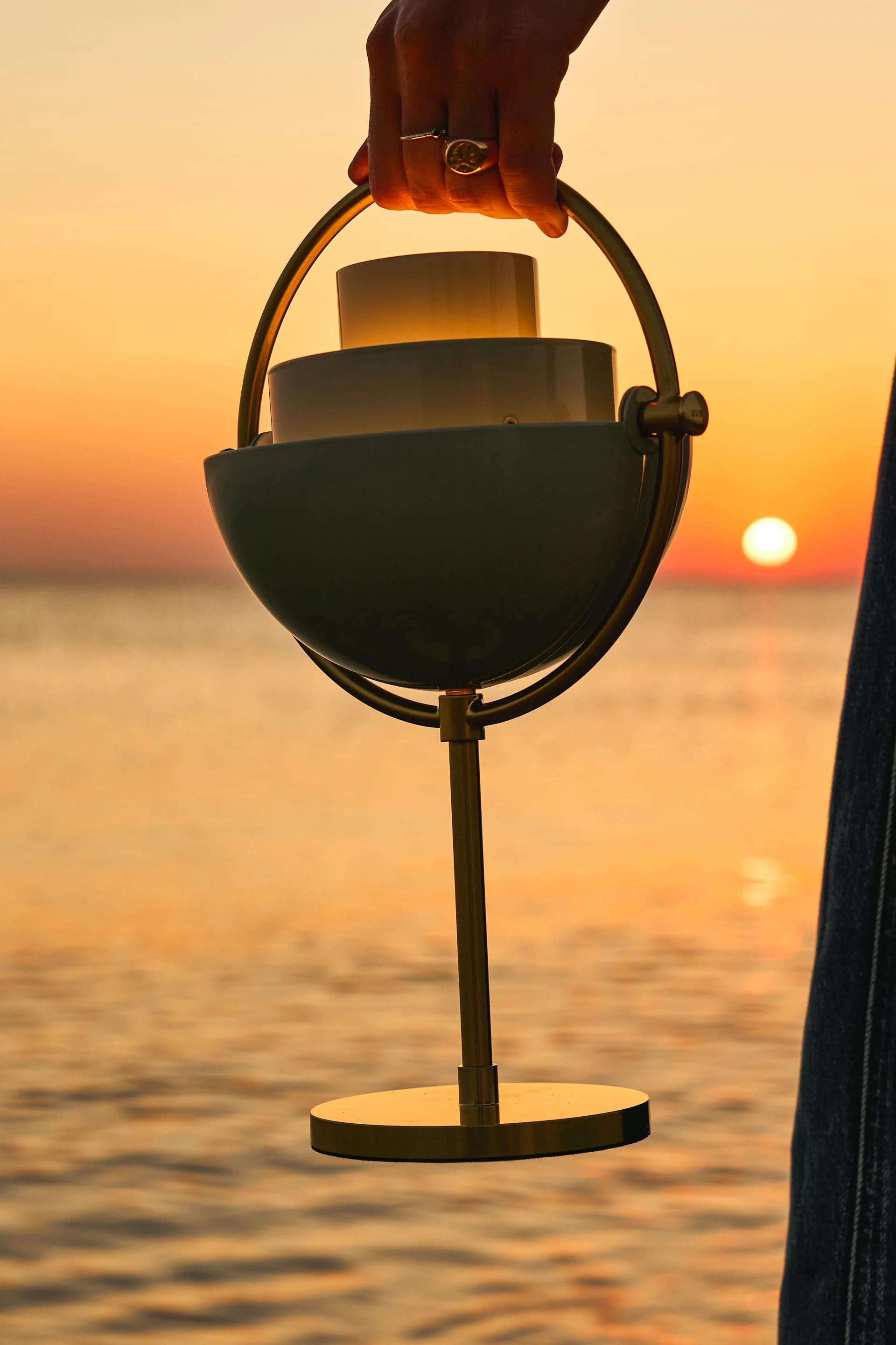 Eine Hand hält eine moderne Akku-Tischleuchte mit einem glühenden Licht im Inneren, vor einem leuchtend orangefarbenen Sonnenuntergang über dem Meer.