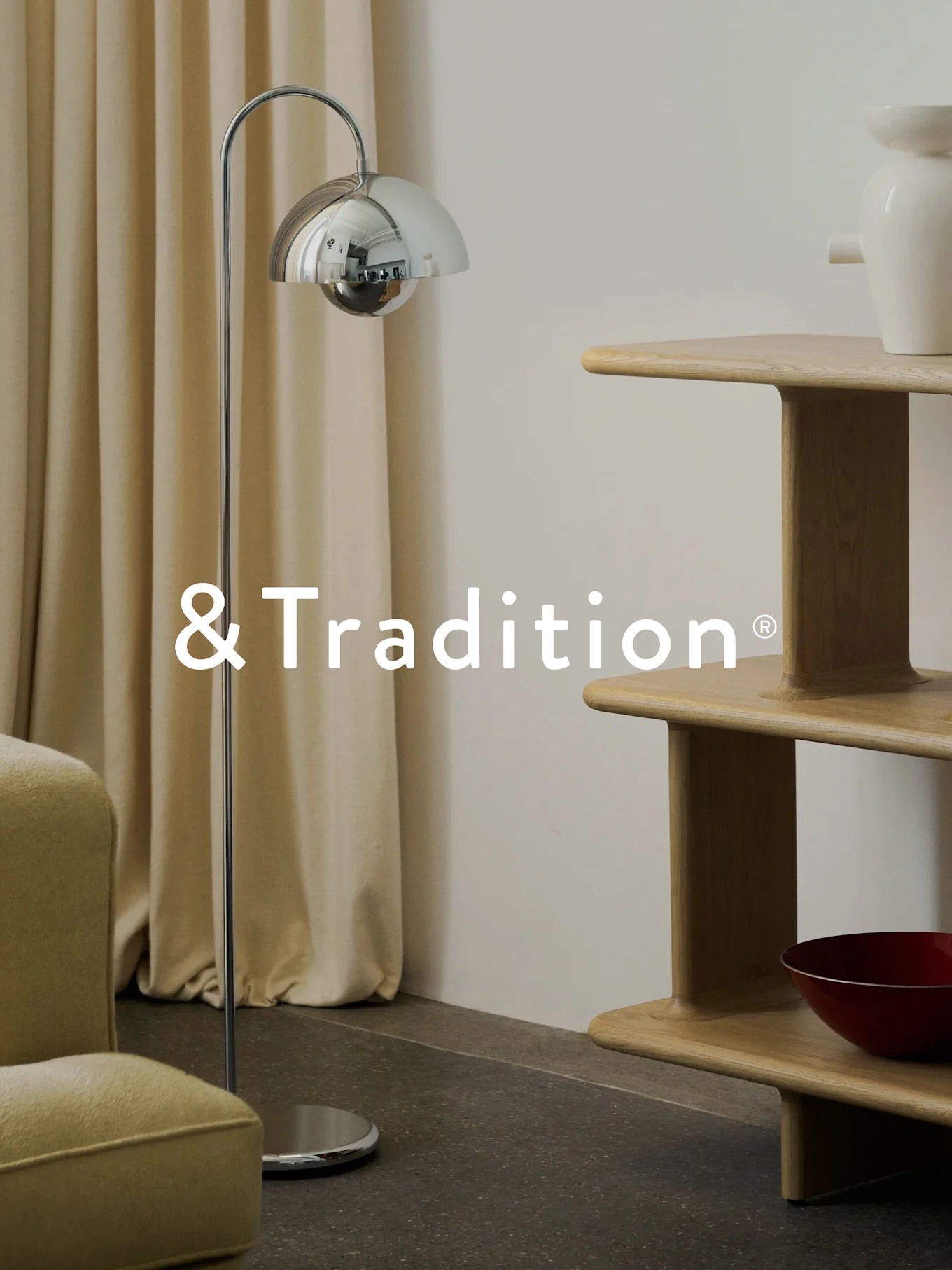 Ein modernes Interieur mit einer Chrom-Stehlampe, einem mehrstufigen Regal aus hellem Holz und einem Sessel. &Tradition® Logo.