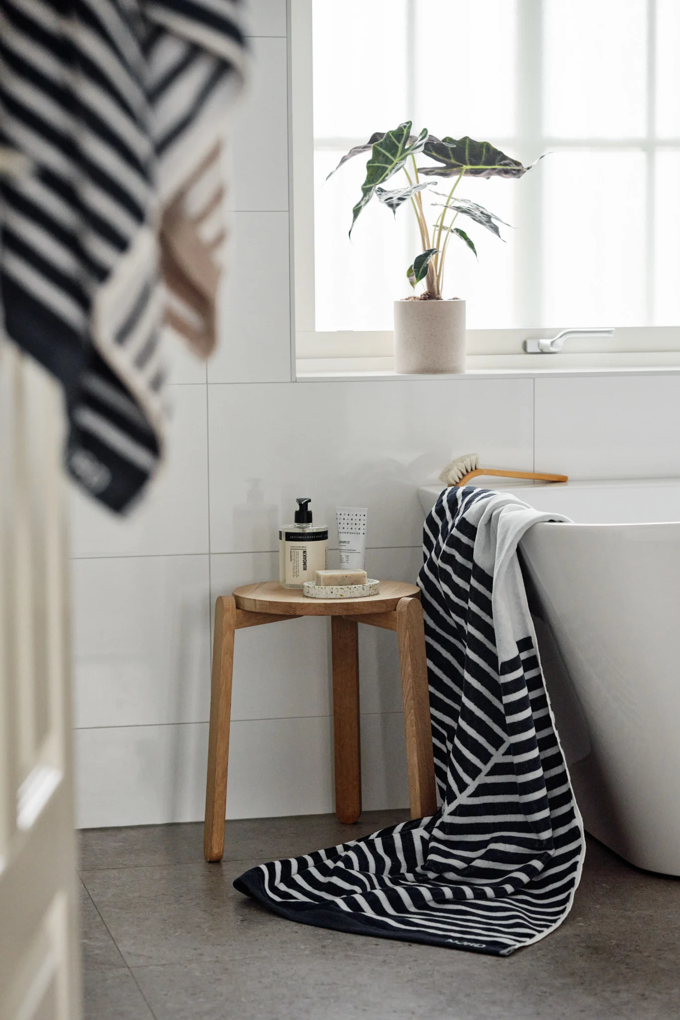Modernes Badezimmer mit schwarz-weiß gestreiften Handtüchern, einem Holzhocker mit Badprodukten und einer Topfpflanze am Fenster.