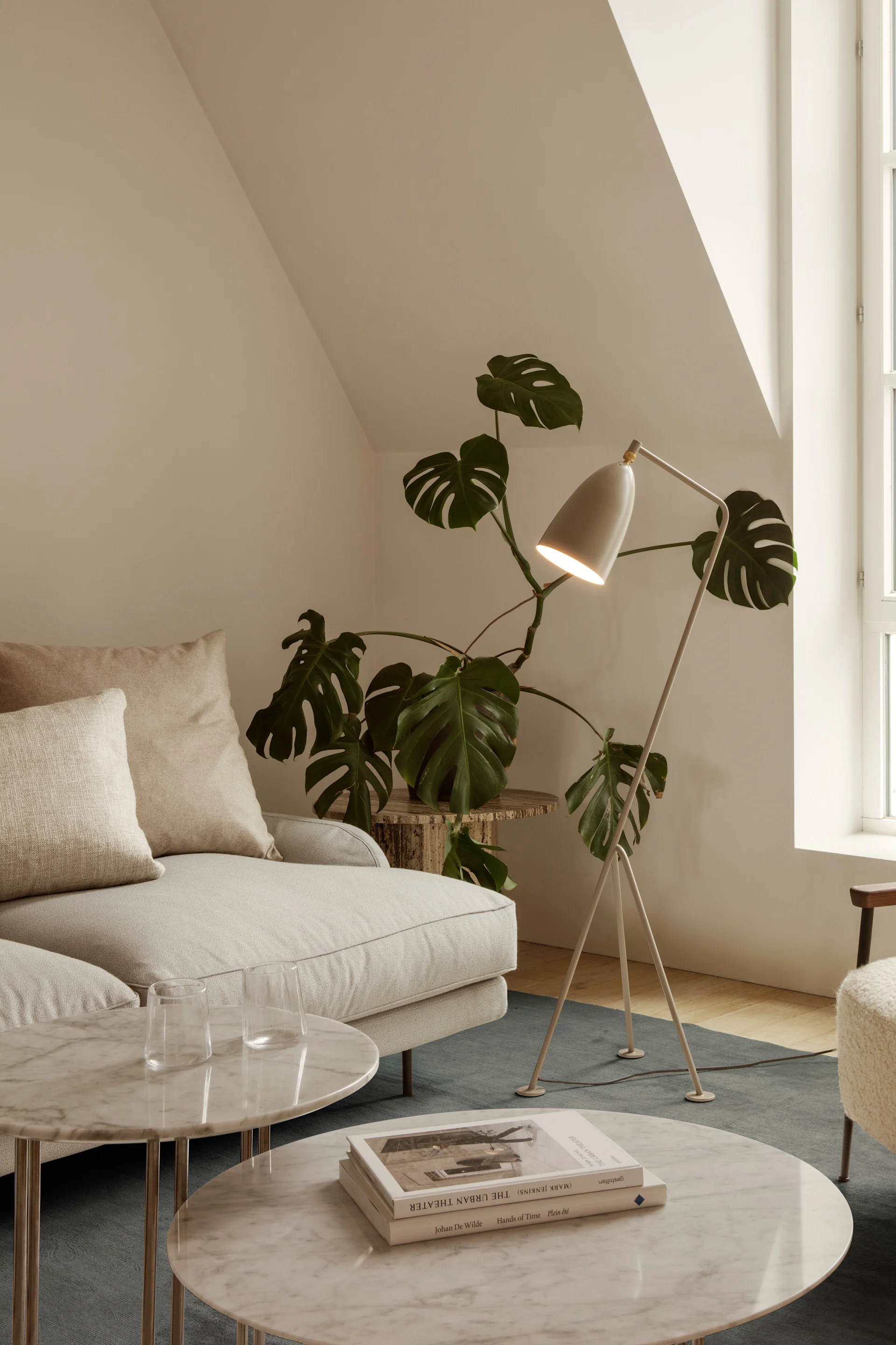 Ein helles Wohnzimmer mit cremefarbenem Sofa, Marmor-Couchtischen, Stehlampe, Pflanze und flauschigem Sessel am Fenster.
