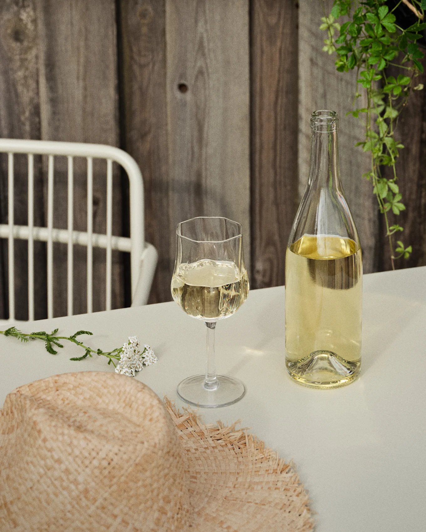 Eine Flasche und ein Glas Weißwein auf einem Tisch mit einem Strohhut und weißen Blumen, vor einer Holzwand und grünen Ranken.