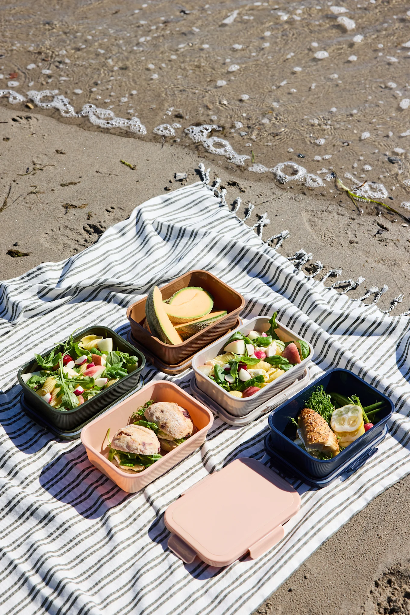 Fünf offene, farbenfrohe Lunchboxen mit Salaten, Sandwiches und Melone, plus eine geschlossene rosa Box, auf einer gestreiften Stranddecke.