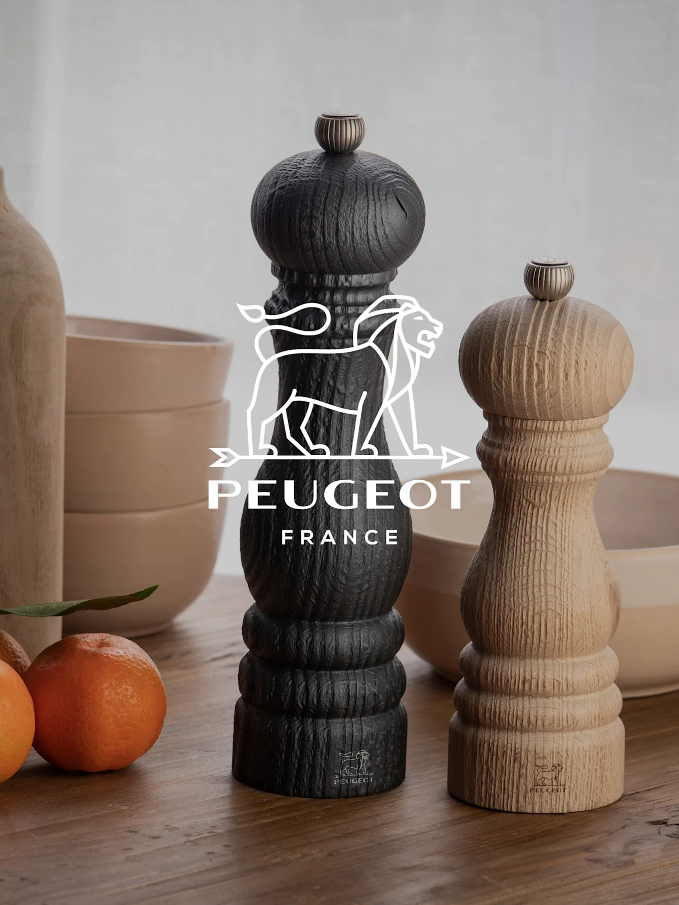 Zwei Peugeot Pfeffermühlen, eine schwarz und eine aus Naturholz, auf einem Holztisch mit Mandarinen und Schalen.