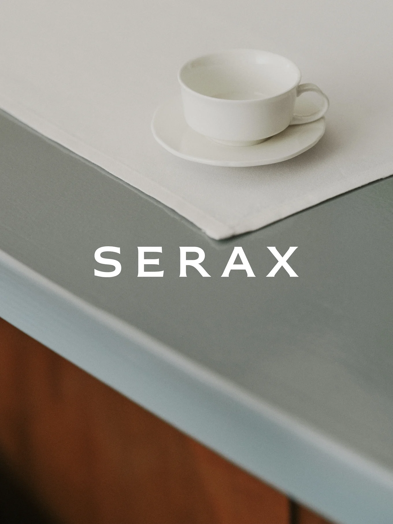 Eine weiße Teetasse und Untertasse auf einem hellen Leinenplatzdeckchen auf einem grauen Tisch mit dem Wort SERAX.