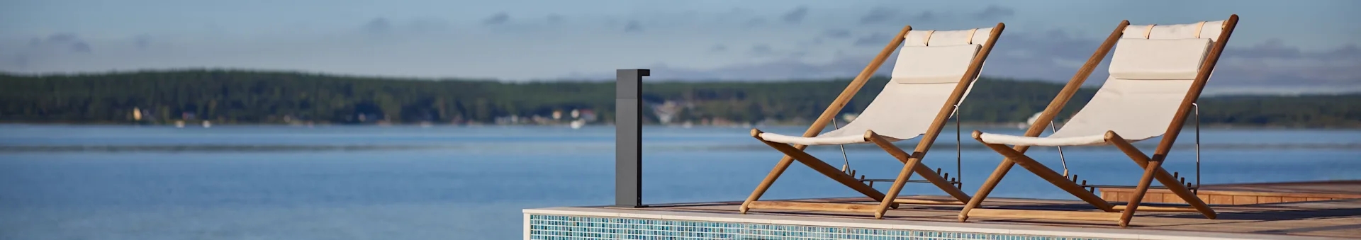 Zwei weiße Stoff- und Holzliegestühle auf einer Terrasse mit Blick auf einen See und einen Wald in der Ferne.