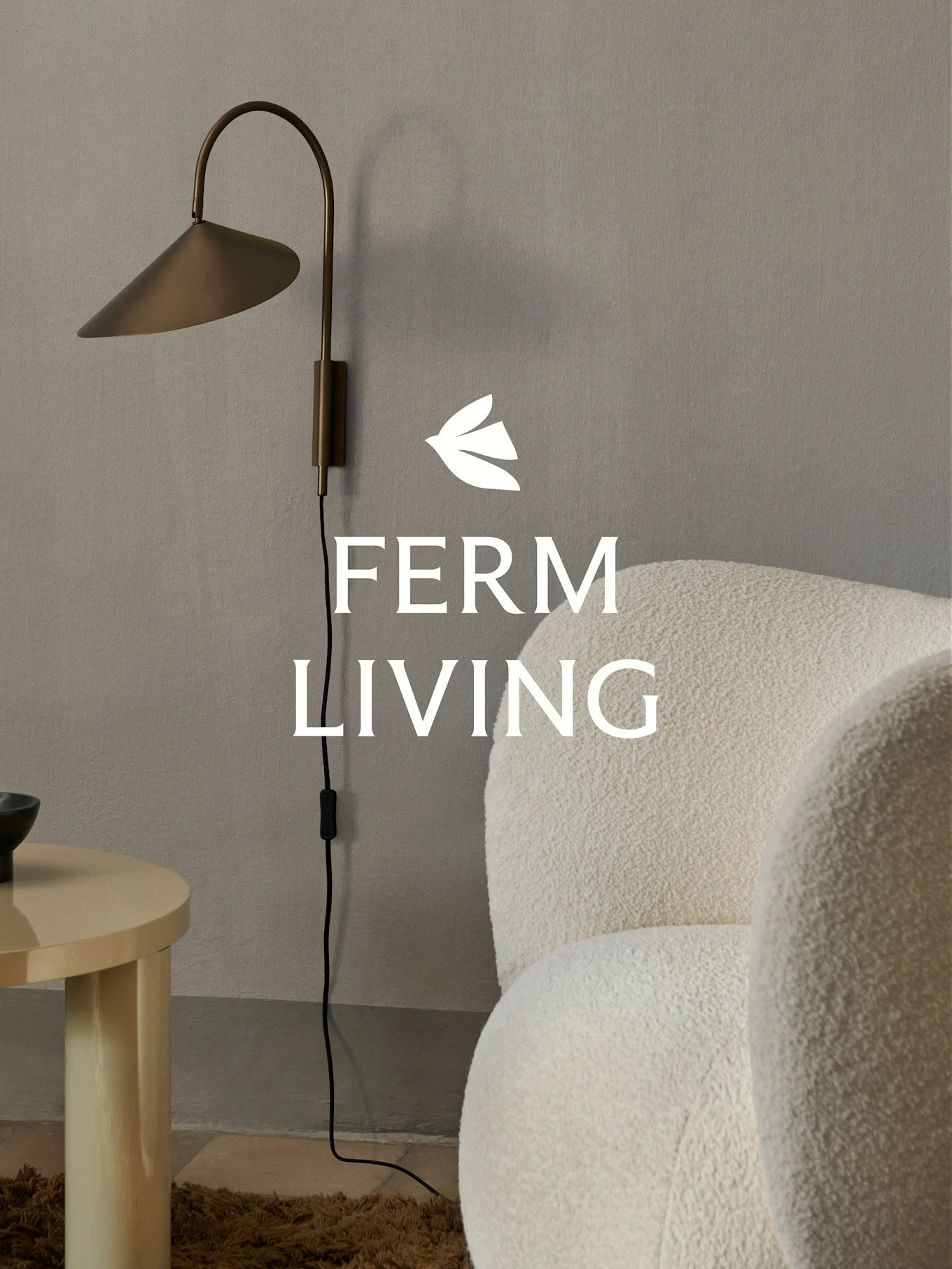 Ferm Living Logo über einer Messing-Wandlampe, einem cremefarbenen Bouclé-Sessel und einem hellen Holz-Beistelltisch in minimalistischer Umgebung.