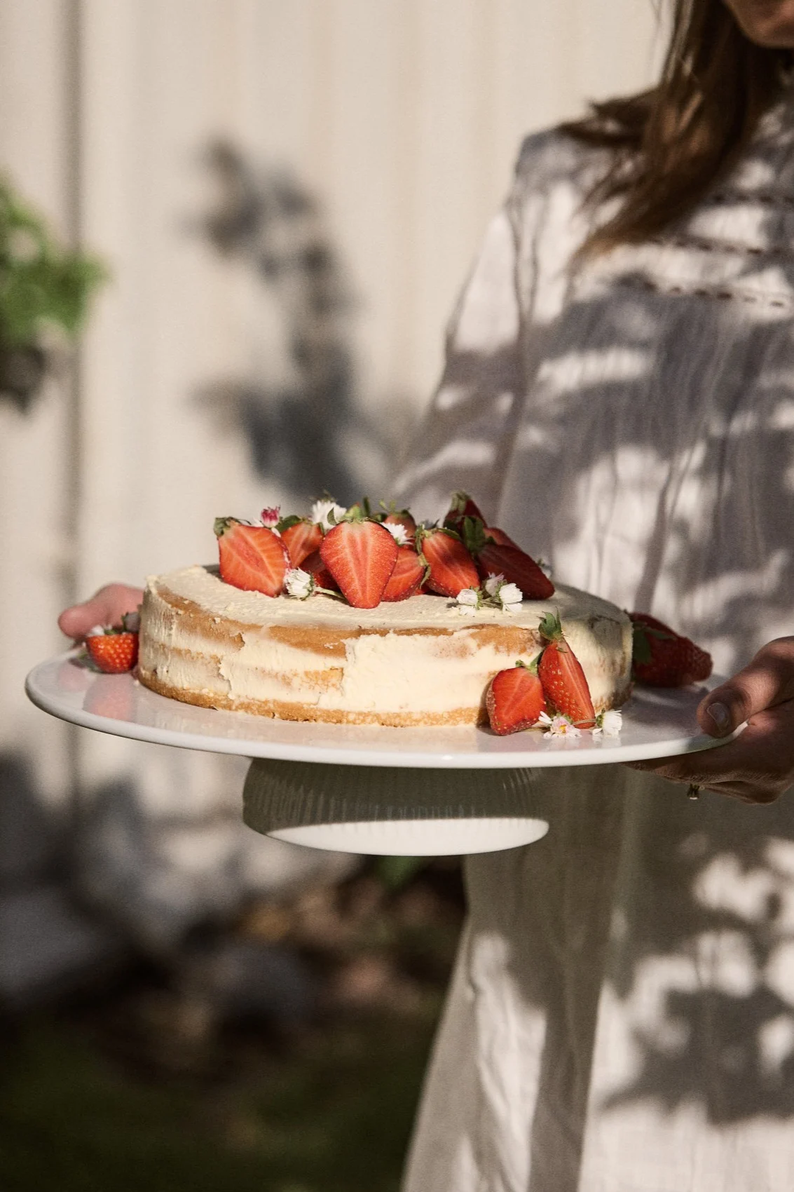 Person hält einen runden Kuchen, der mit frischen halbierten Erdbeeren und kleinen weißen Blüten belegt ist, auf einer weißen Kuchenplatte im Freien.