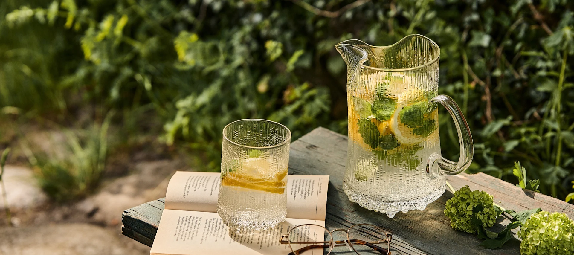 Strukturierte Glaskanne und Becher mit Zitronen- und Minzwasser, ein offenes Buch und Brillen auf einer Holzoberfläche im Freien.