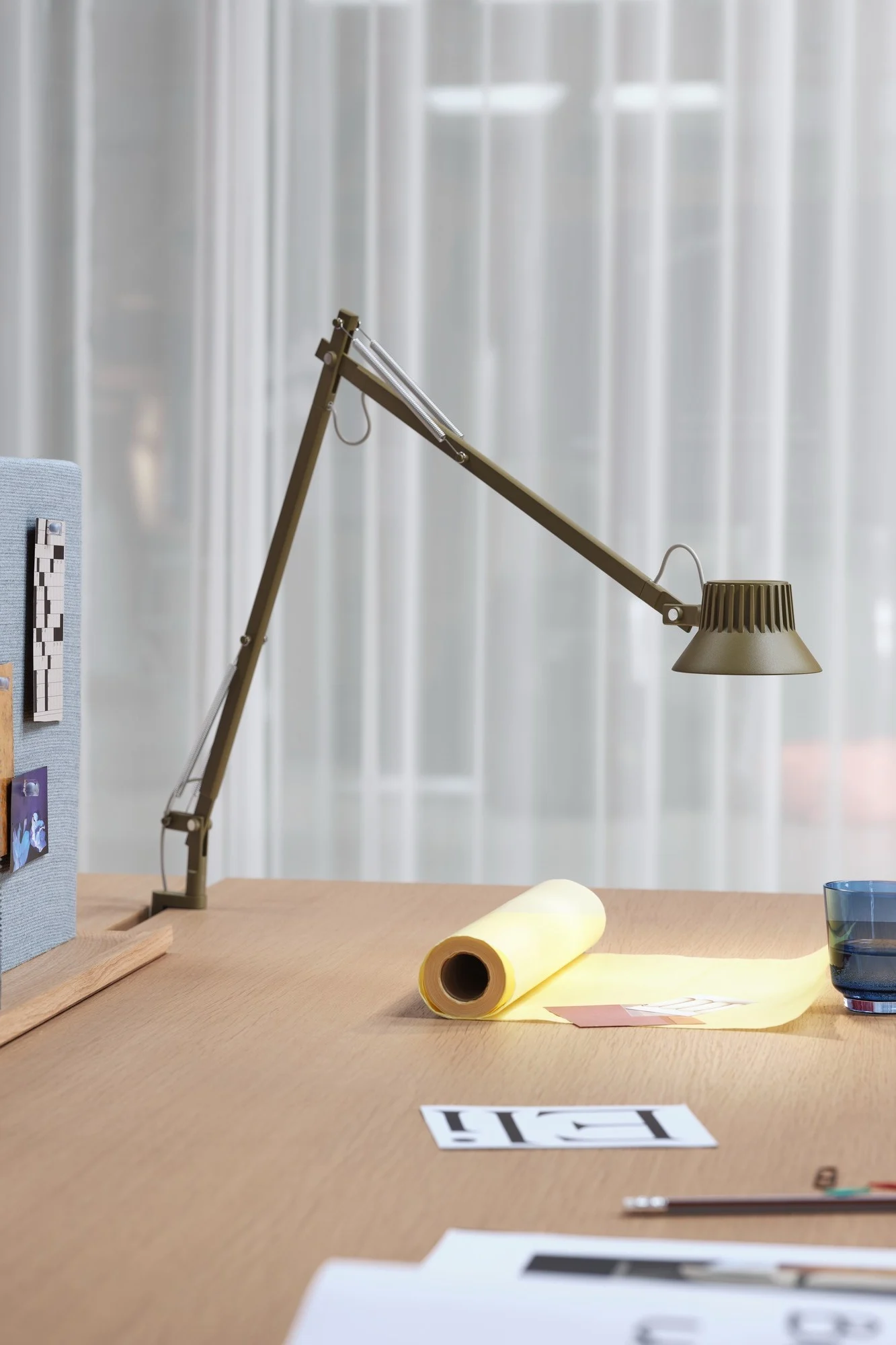 Eine olivgrüne Gelenkschreibtischlampe beleuchtet einen hellen Holzschreibtisch mit gelbem Papier, einem blauen Glas und Dokumenten.