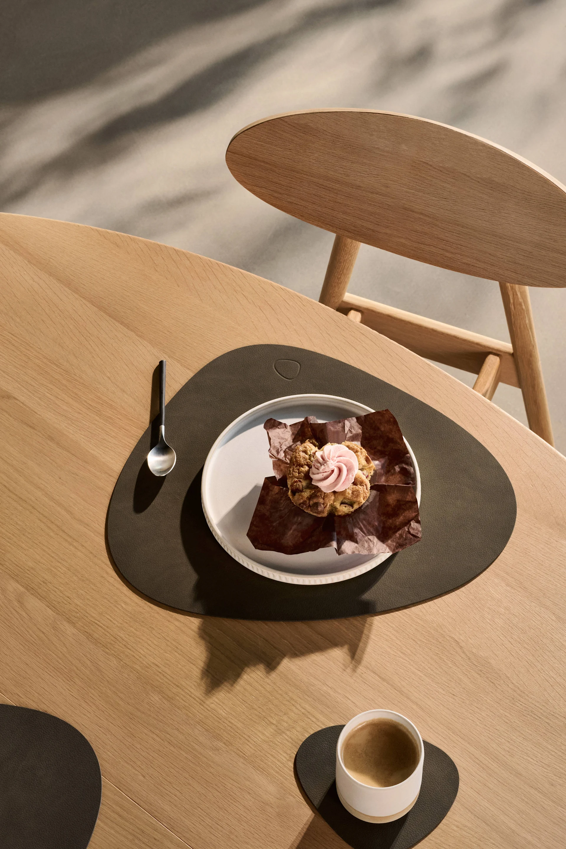Muffin mit rosa Zuckerguss und Kaffee auf Holztisch mit dunklen Platzsets und Untersetzern.