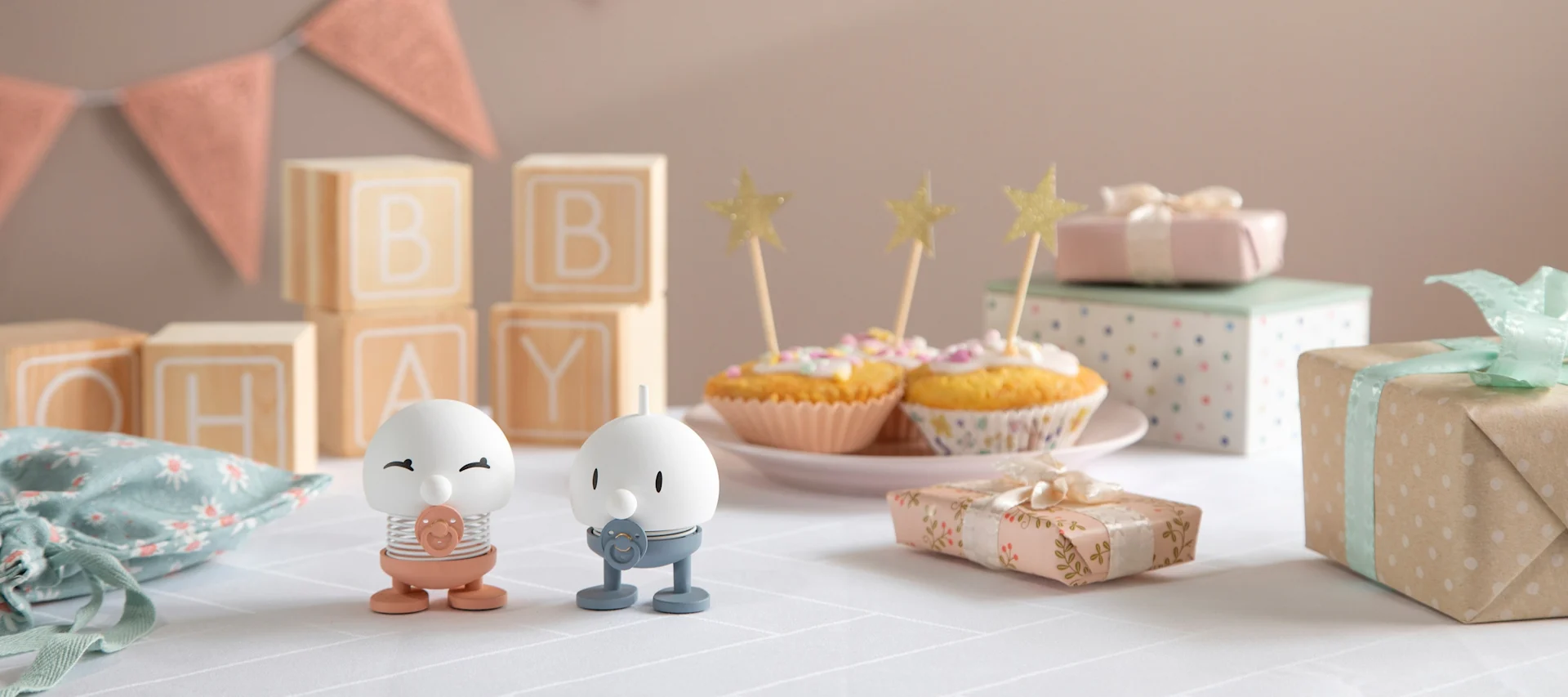 Die Hoptimisten Baby Bimble sind in Rosa und Blau erhältlich und stehen auf einem mit Geschenken, Girlanden und Muffins dekorierten Tisch.
