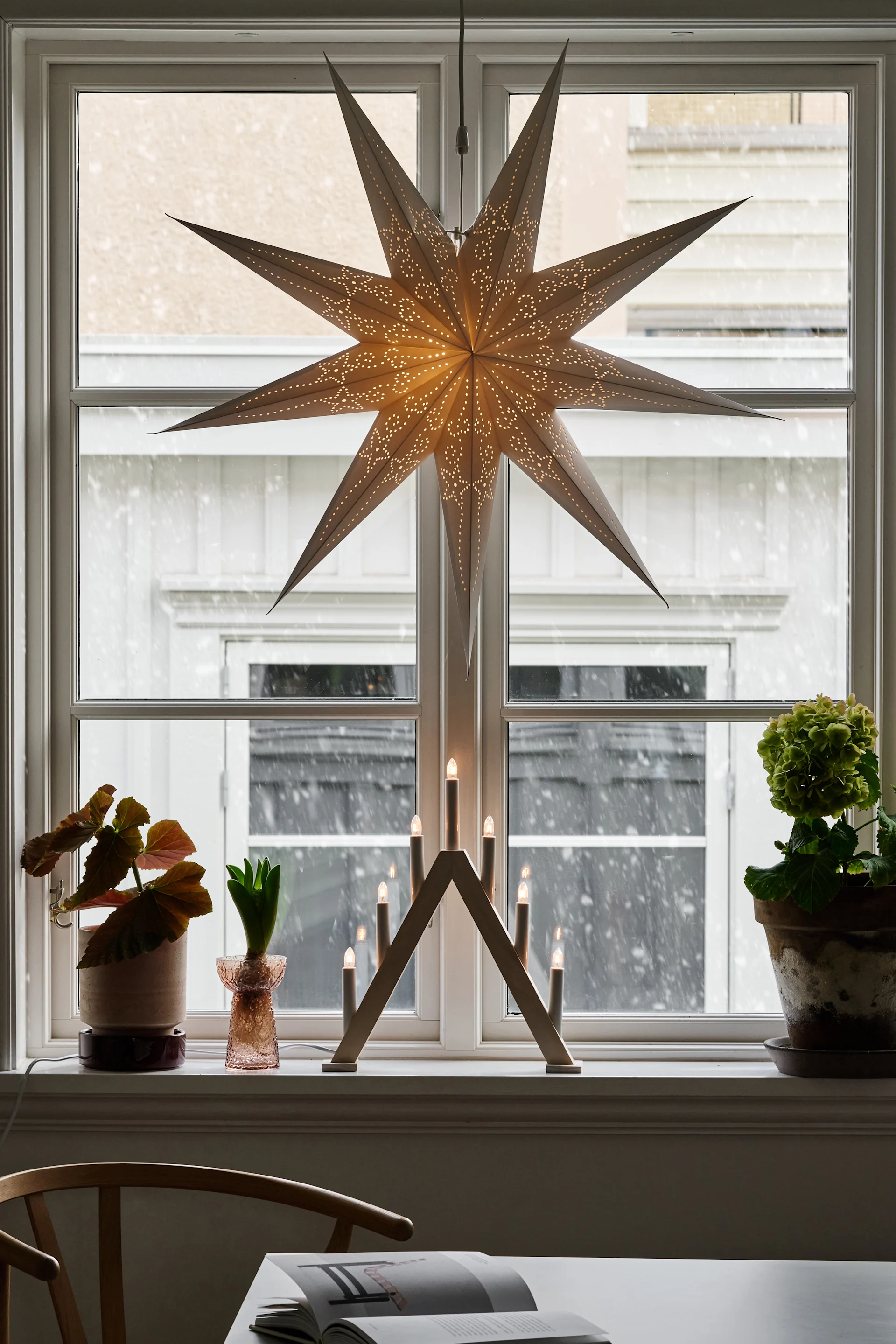 Eine weiße plissierte Pendelleuchte hängt in einem Fenster, umgeben von transparenten Vorhängen, über einem Heizkörper und einer Schale mit einer Kugel.